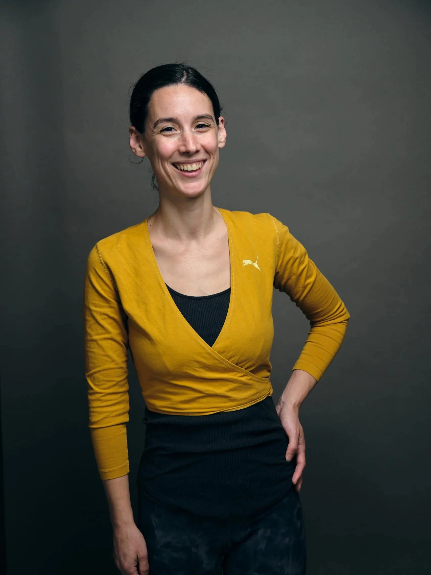 A woman with dark hair, smiling, wearing a yellow Adidas sweater over a black top, and black pants, standing against a gray background.