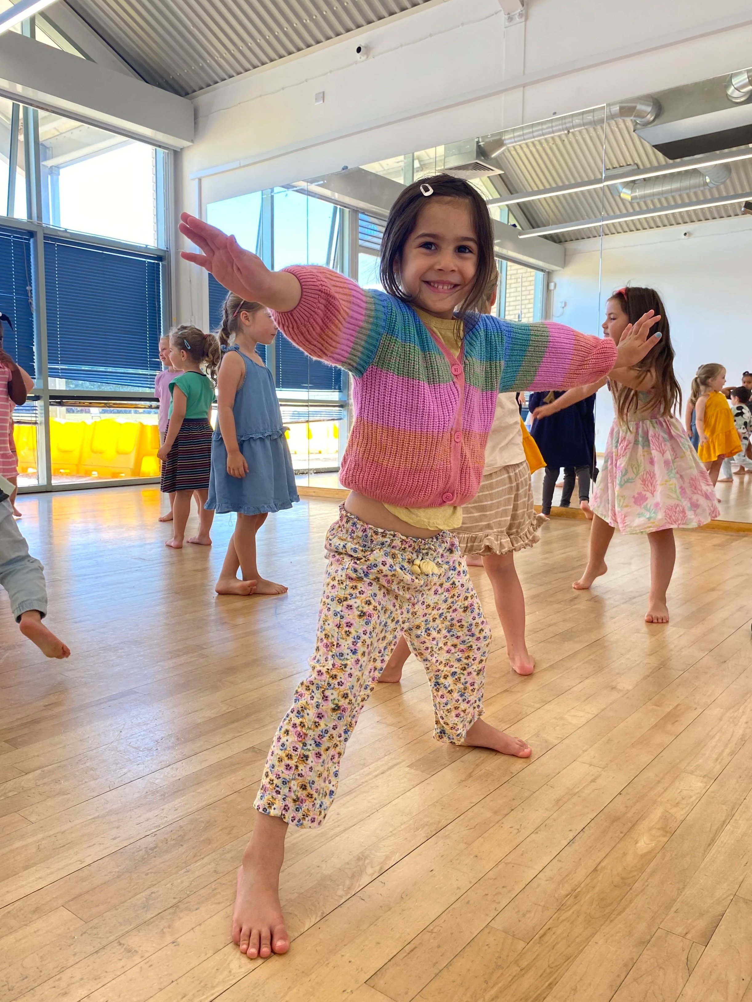 Young girl smiling with arms outstretched in a dance studio filled with children.