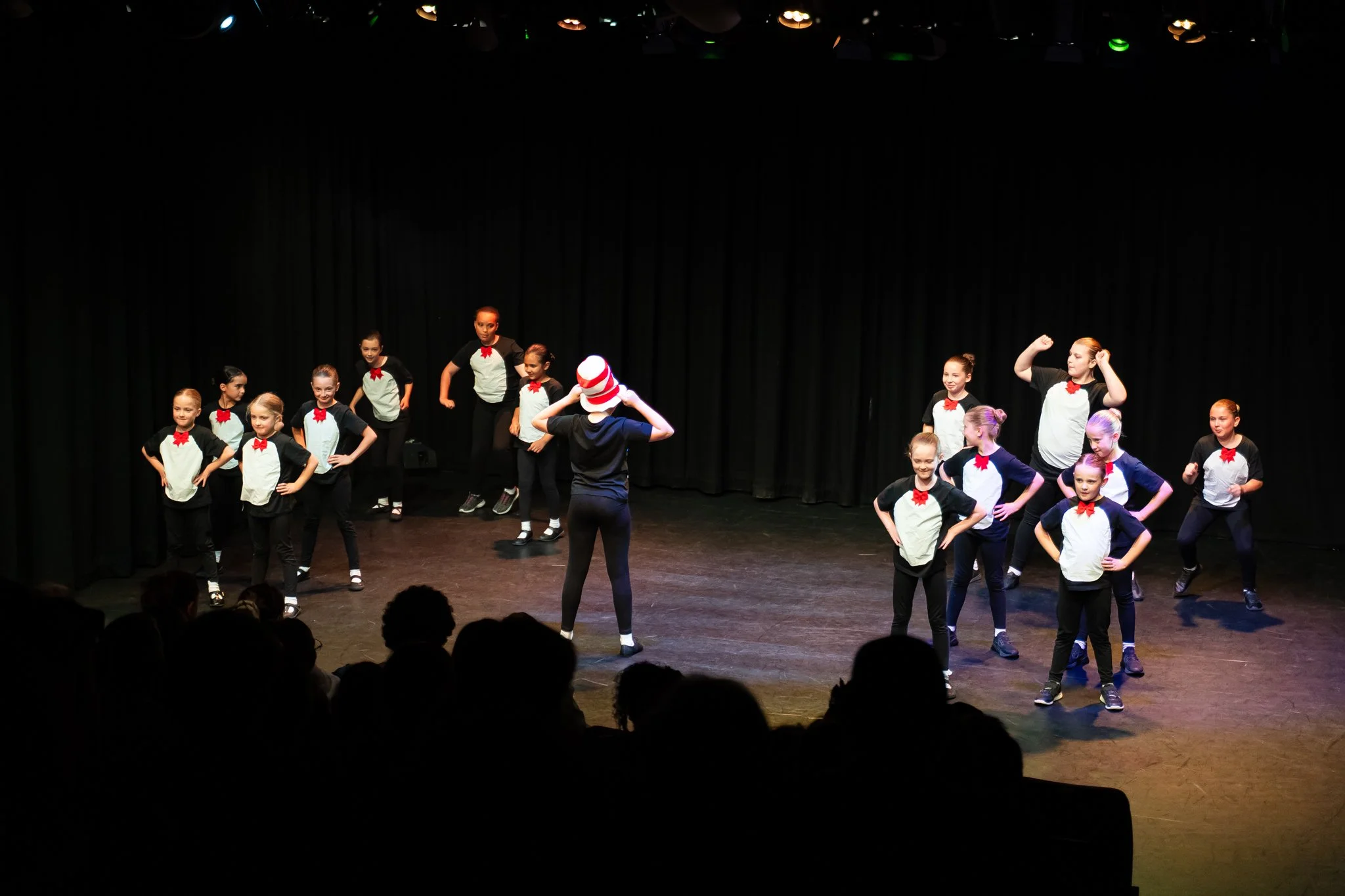 Children performing on stage in a dance recital, wearing matching black and white costumes with red bow ties, under colorful stage lighting.