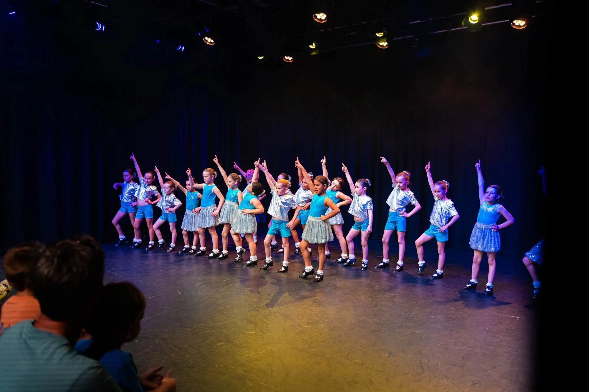 Group of young girls performing a dance on stage in a recital, dressed in coordinated costumes, with some raising their arms and others standing with hands on hips, under colorful stage lights.