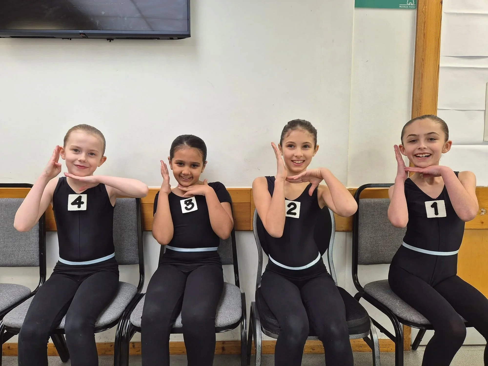 Four young girls sitting on chairs against a wall, posing with their hands near their faces, each wearing a black dance outfit with a number pinned to their chest, numbered 4, 3, 2, and 1 from left to right.