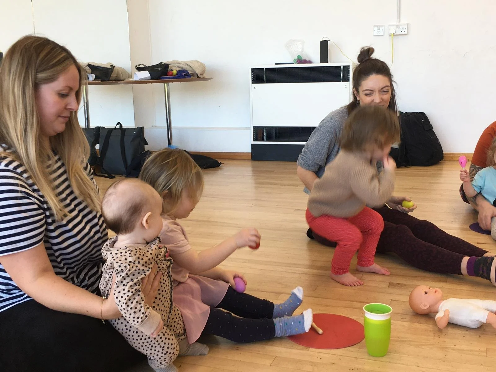 Adults and children sitting on a wooden floor in a room, engaging in a play activity. The children are holding colorful toys, and a doll is lying on the floor nearby. A table with bags and miscellaneous items is in the background.