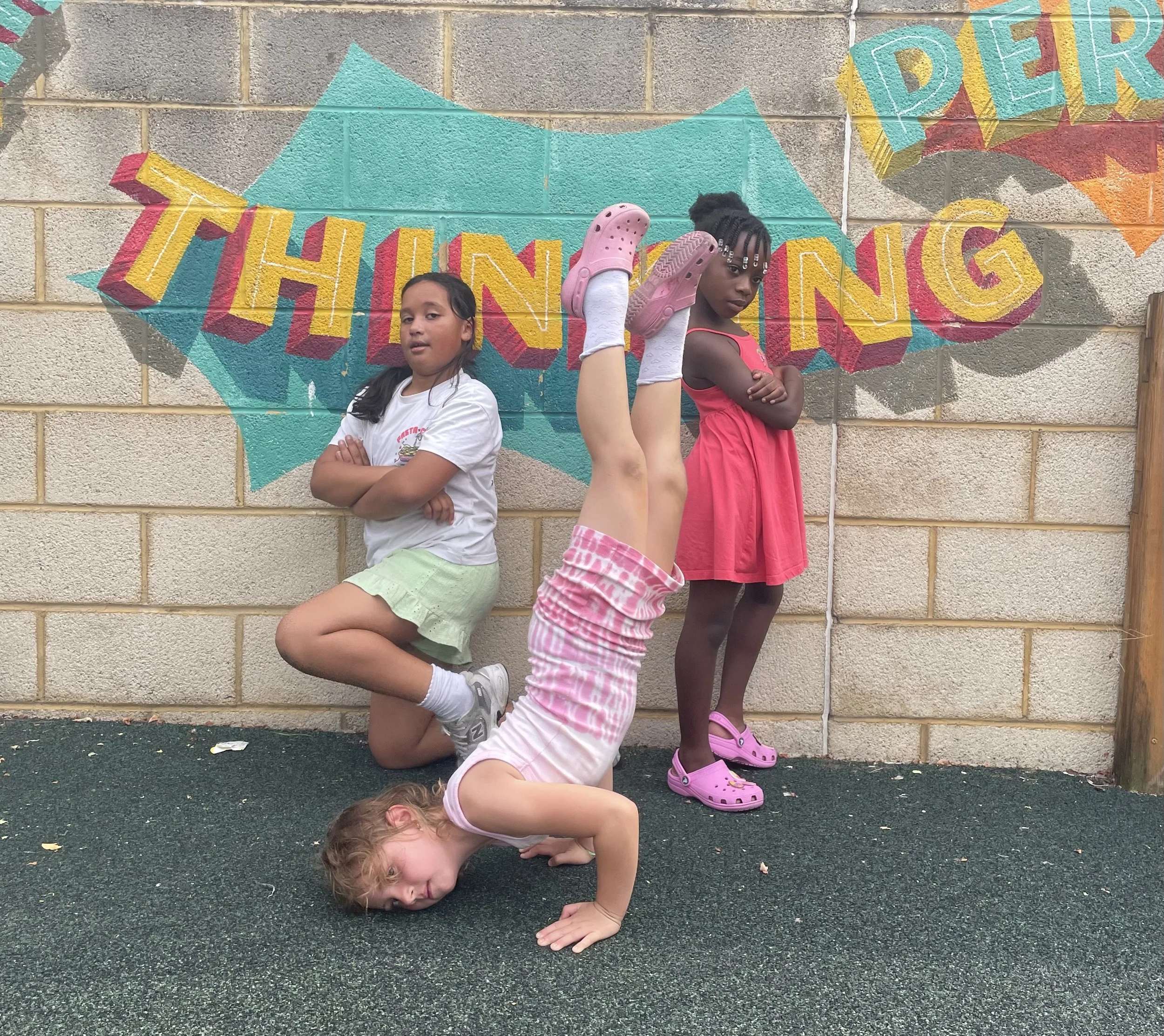 Four children posing in front of a colorful mural that says 'THINKING' on a brick wall. One girl is standing with crossed arms, another girl is kneeling, a third girl is bent backwards doing a handstand, and the fourth girl is standing with her arms crossed, all smiling or appearing playful.