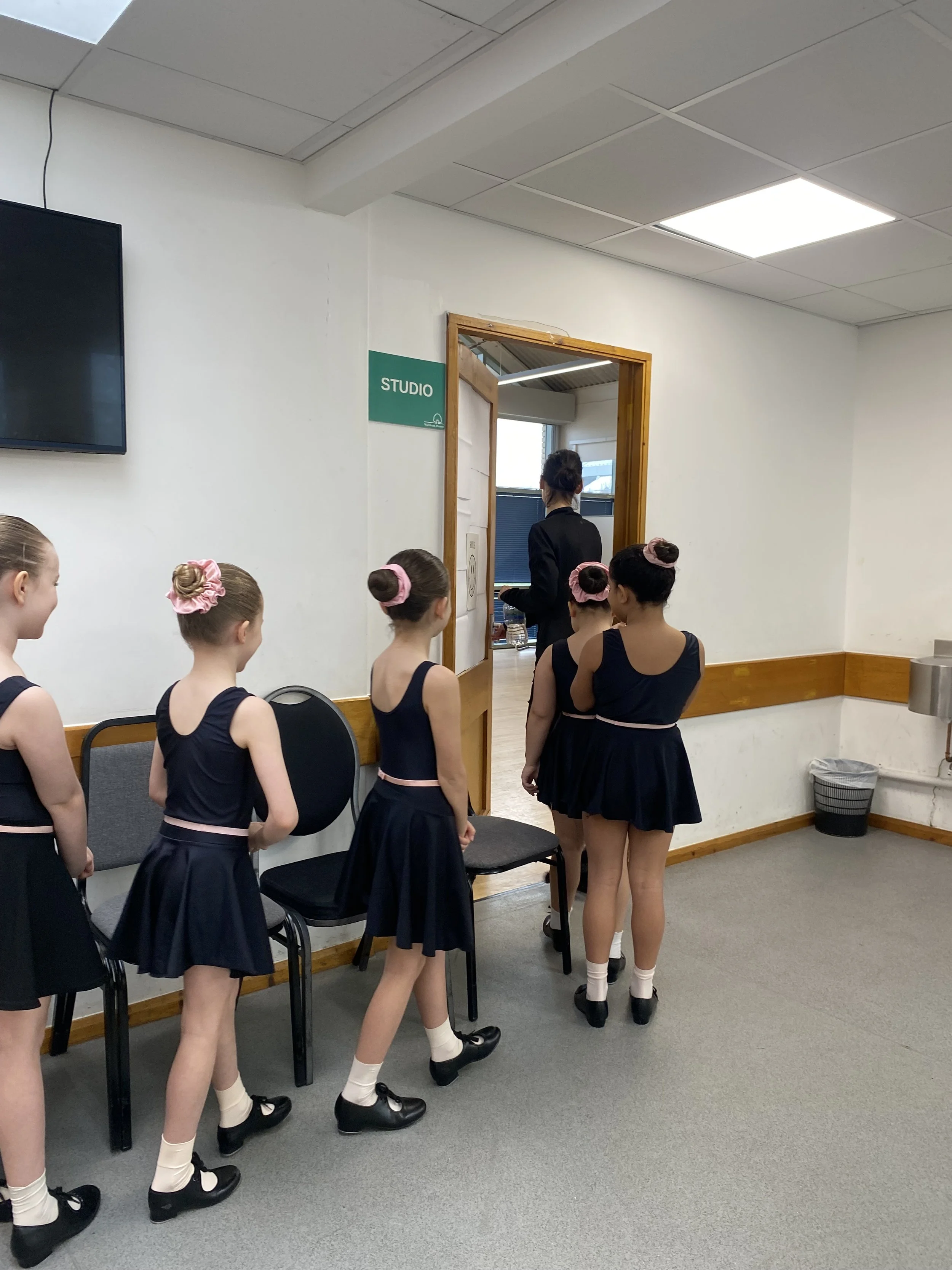 A group of young girls in dance costumes waiting in line outside a studio door, with one girl talking to an instructor inside the room.