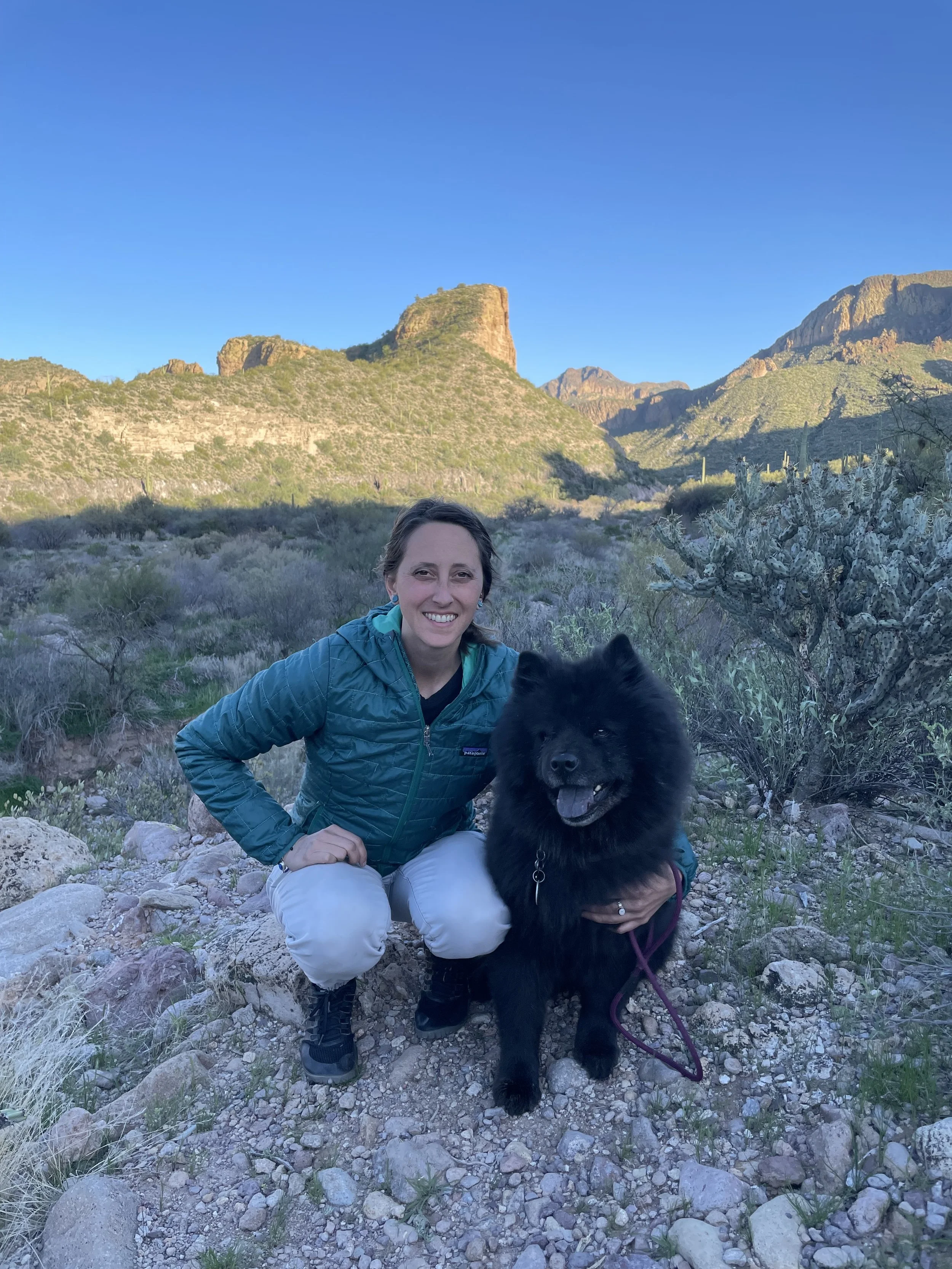A woman smiling and sitting on rocky ground in a desert landscape with a black dog. The background shows mountains, cacti, and a clear blue sky.