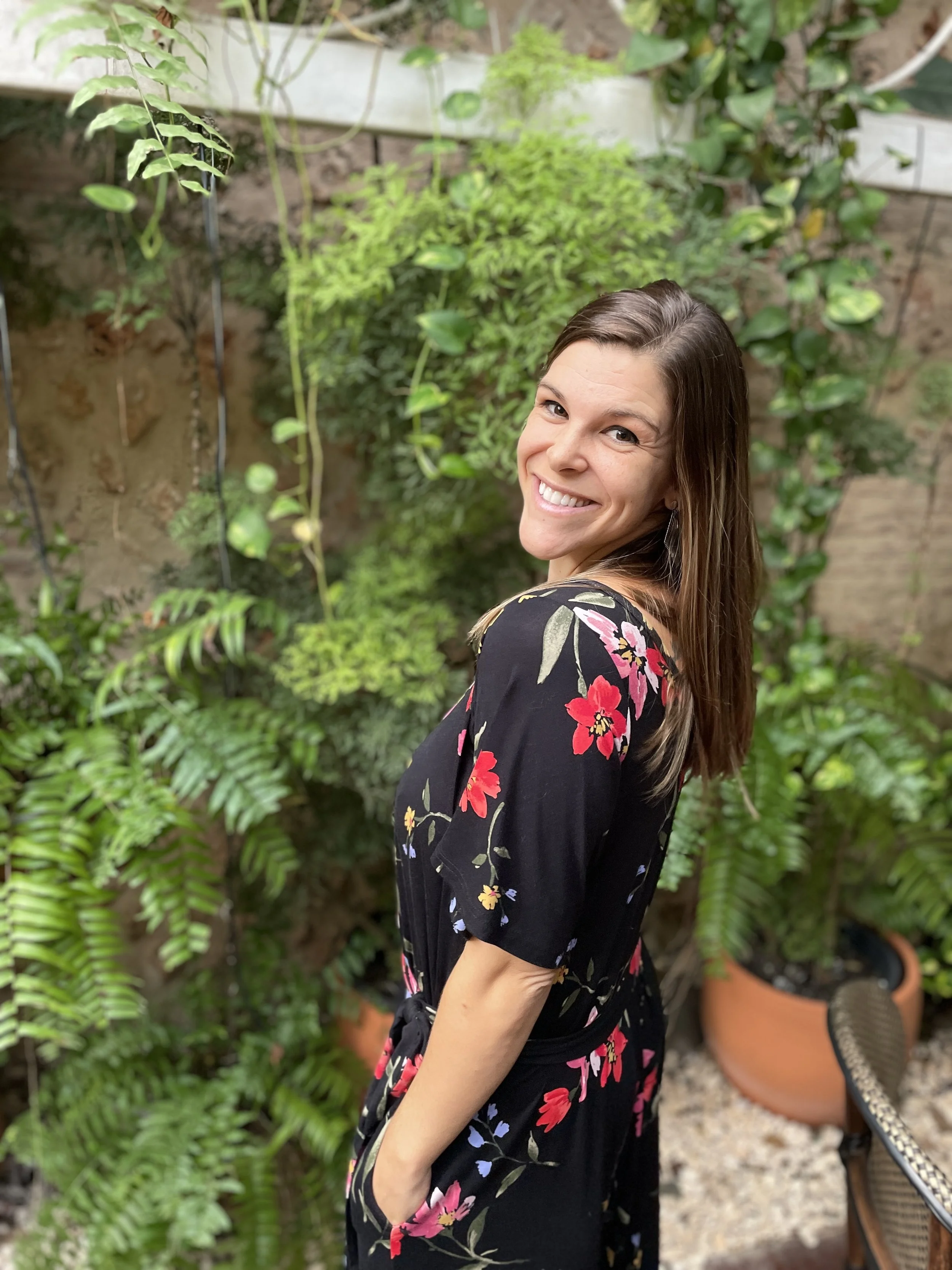 A smiling woman with brown hair in a black floral dress stands outdoors in a garden with lush green plants and potted foliage.