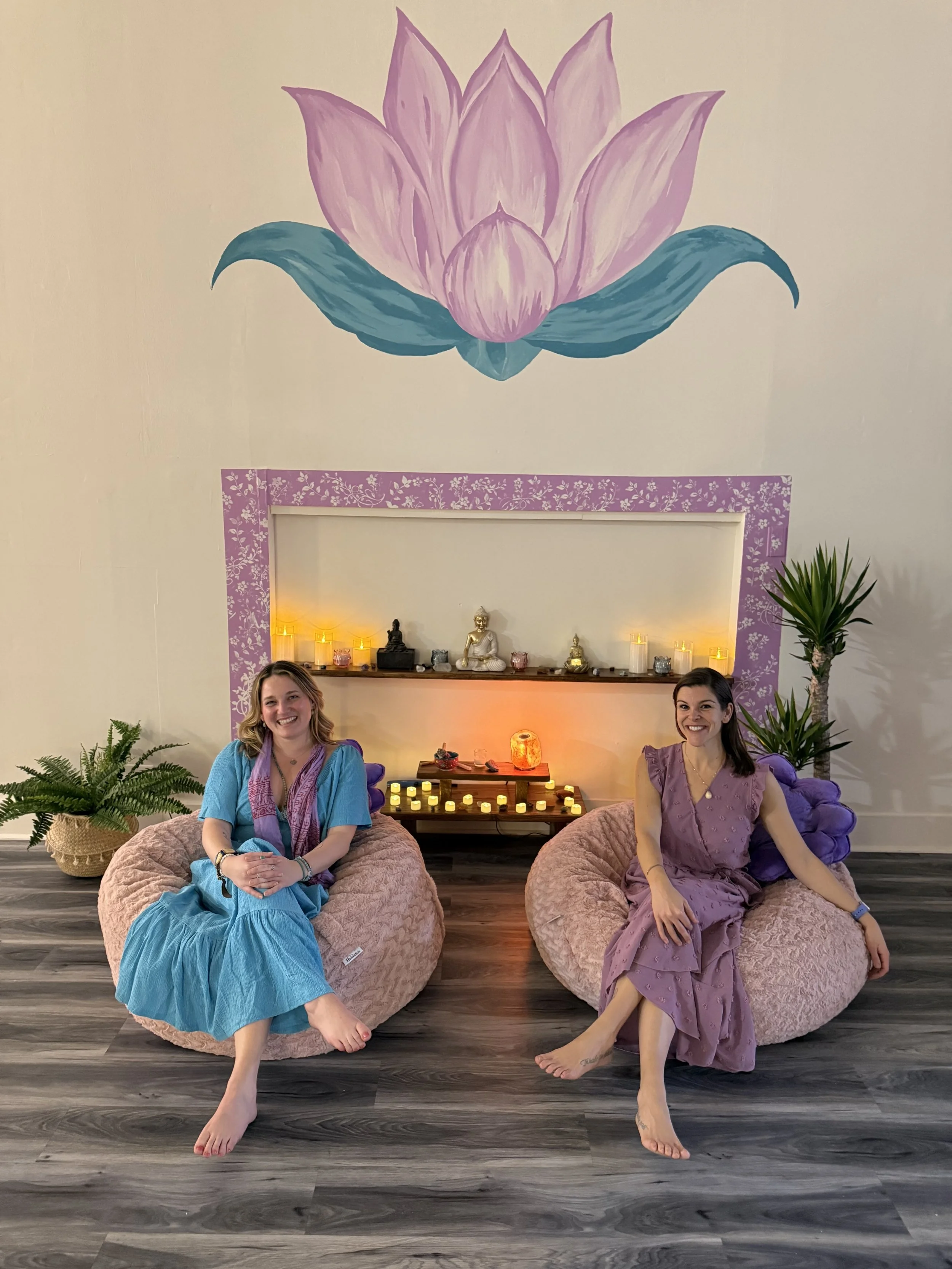 Two women sit on pink bean bag chairs in a yoga or meditation space with candles and small statues on a shelf behind them, and a large painted lotus flower on the wall above.