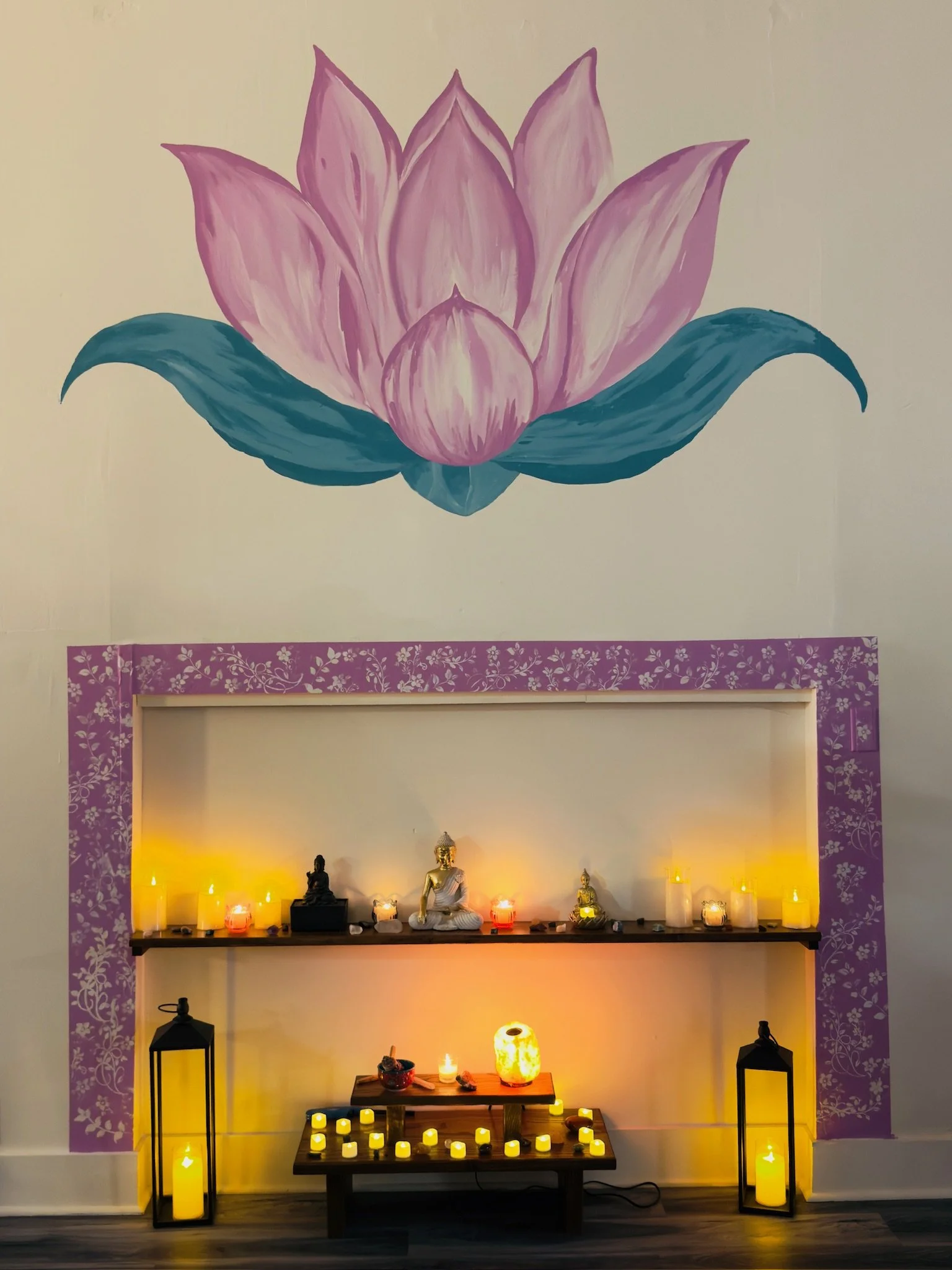 Decorative shrine on a shelf with candles and Buddha statues, curtains, and a large pink and purple lotus flower mural on the wall.