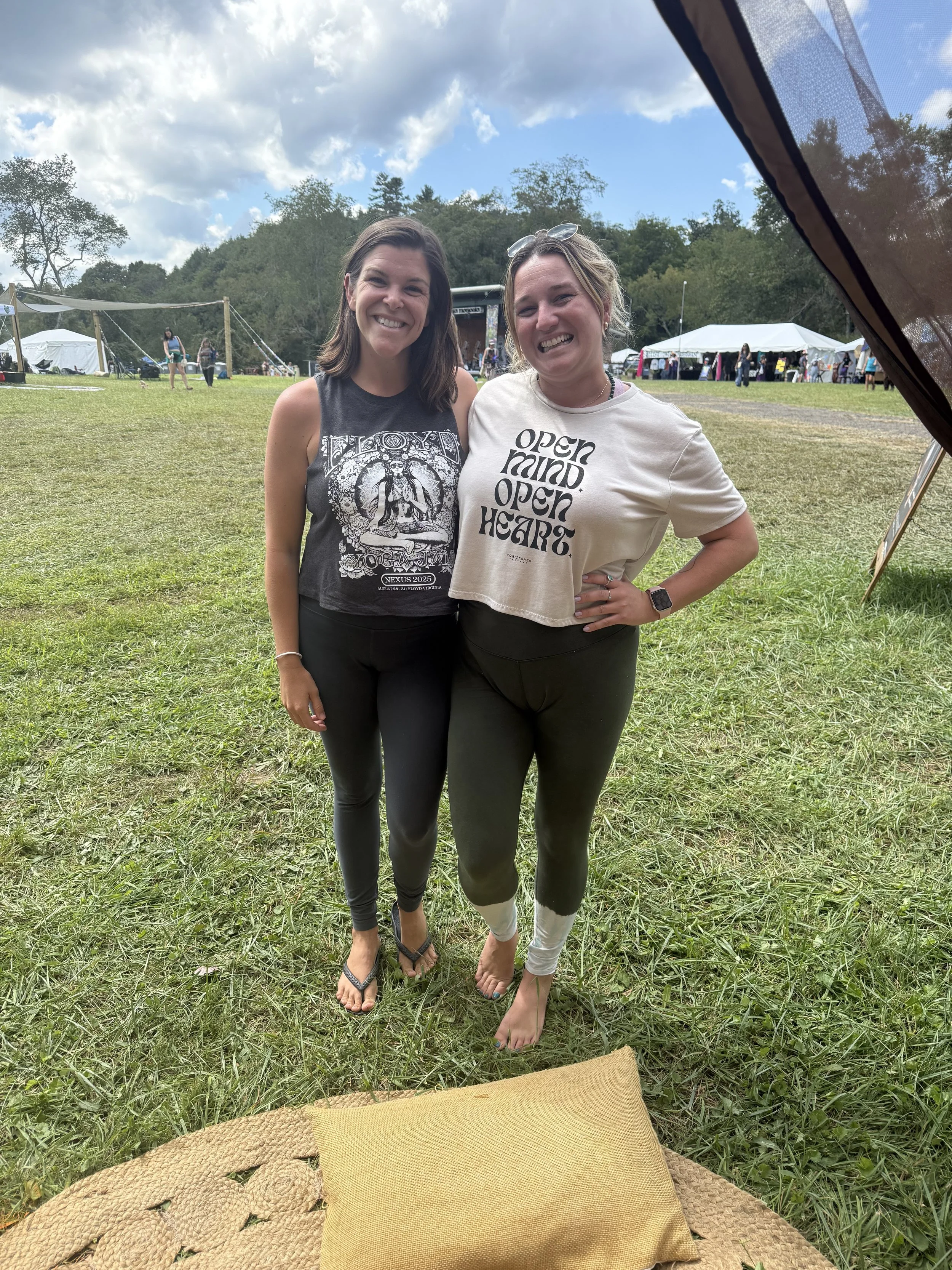 Two women standing together outdoors at a festival on a grassy field, smiling at the camera, with tents and a stage in the background.