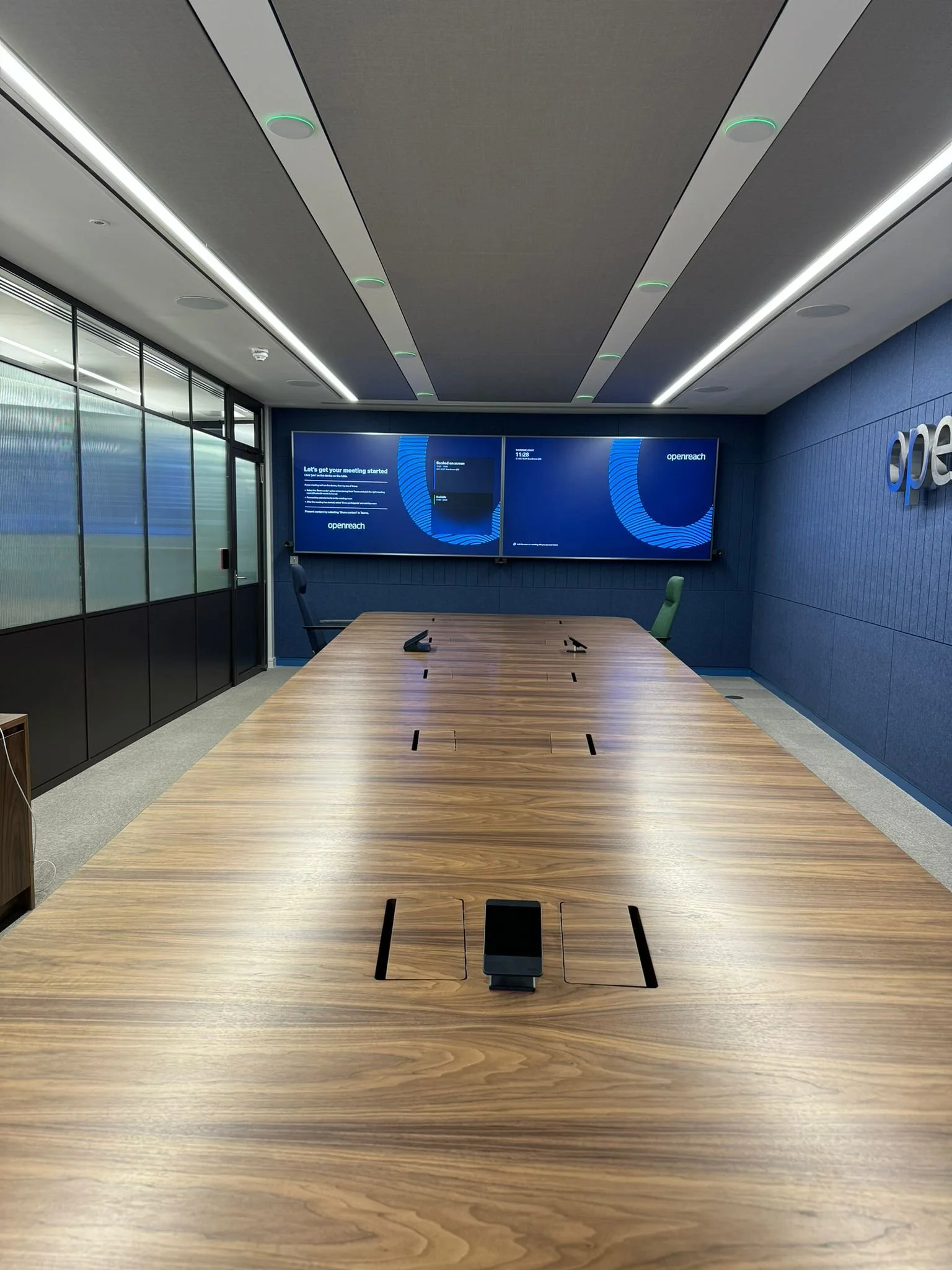 Empty conference room with a large wooden table, chairs, and multiple screens on the wall displaying a presentation.
