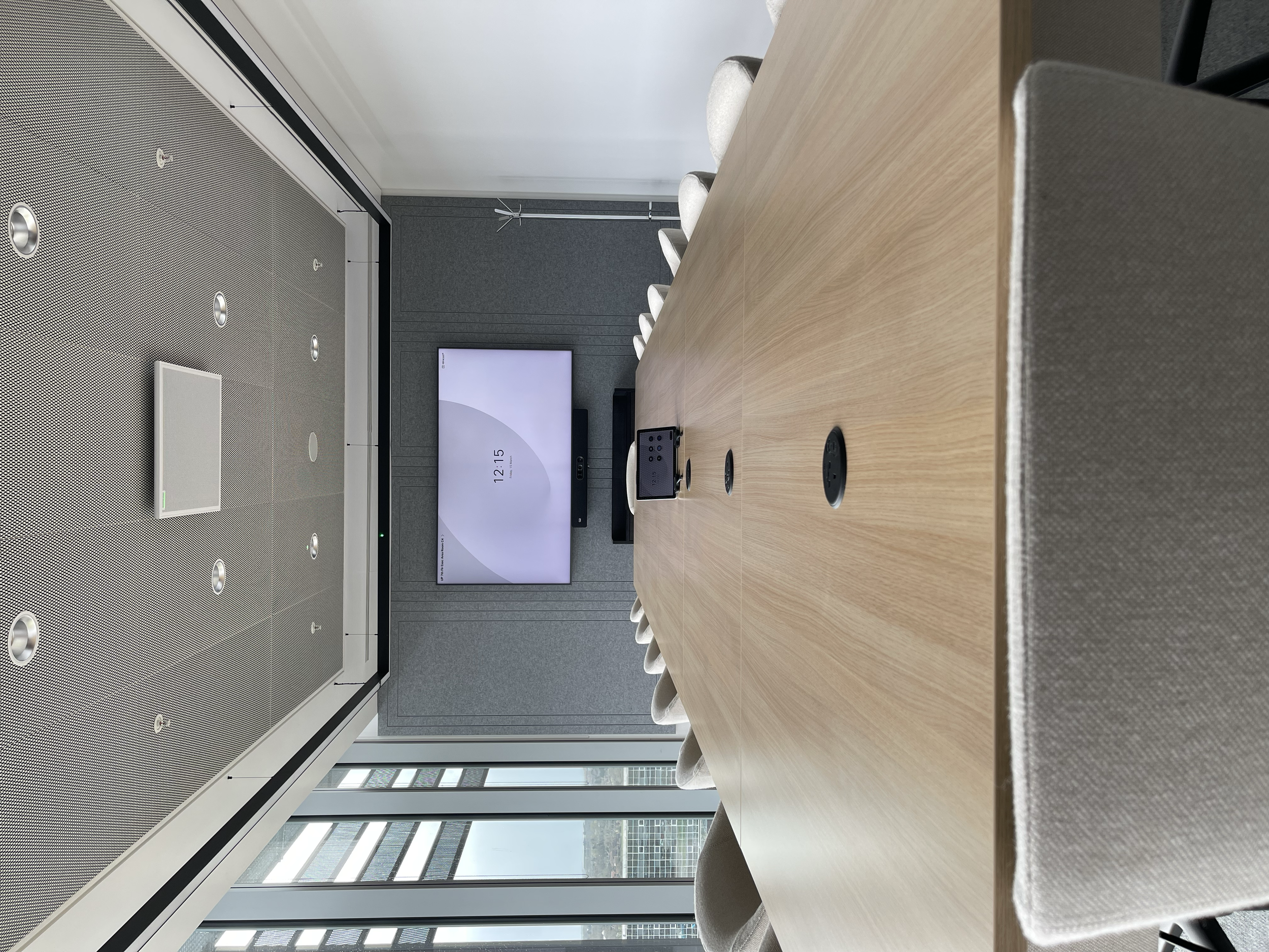 Modern conference room with a long wooden table surrounded by gray upholstered chairs. A large flat-screen TV is mounted on a gray section wall at the far end, with a video conference camera in the center of the table. The room has a ceiling with rec