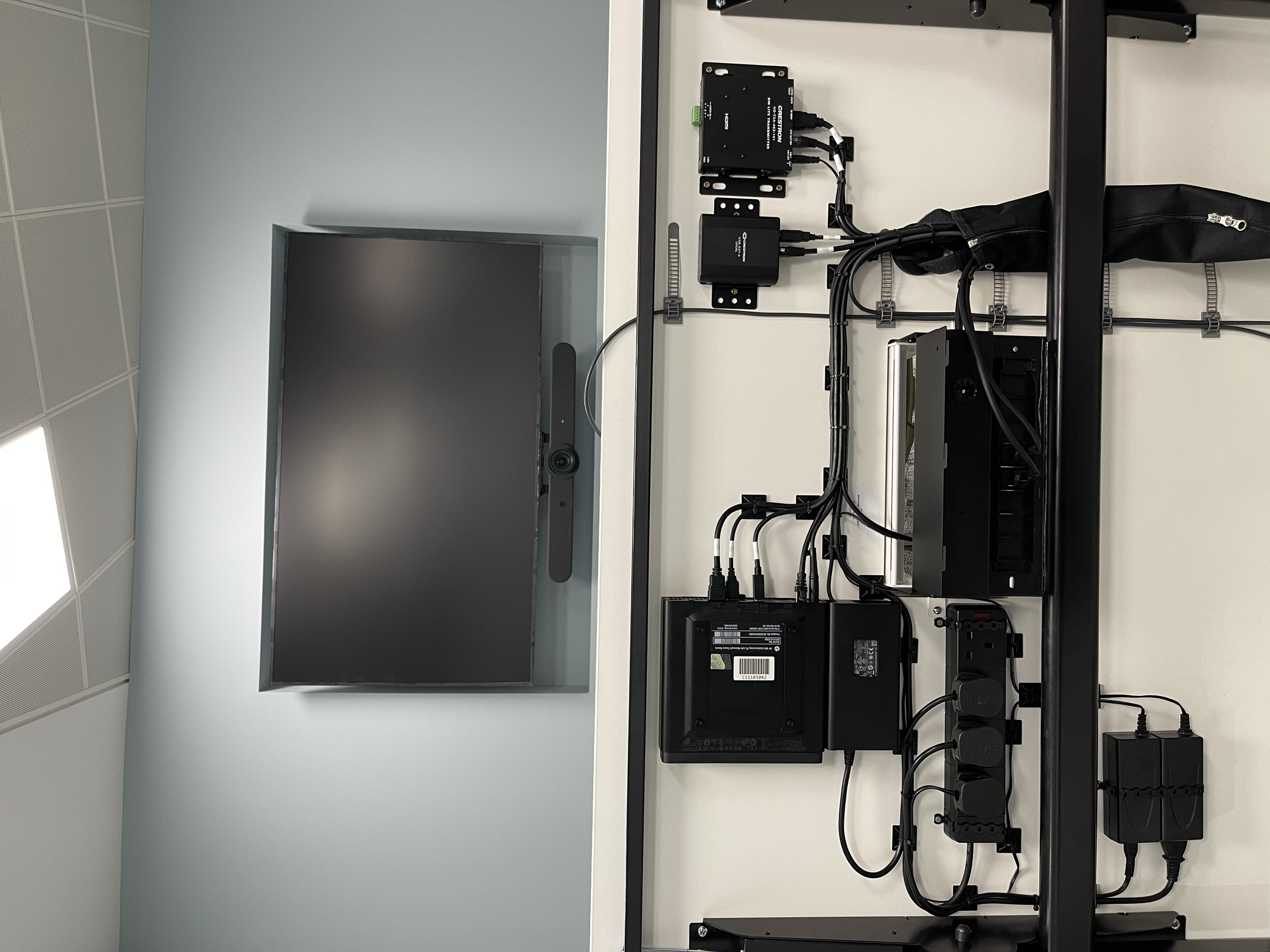 A large flat-screen monitor mounted on a light blue wall and a complex black electronic setup with various devices, cables, and power strips on a white desk, all organized along a black metal frame.