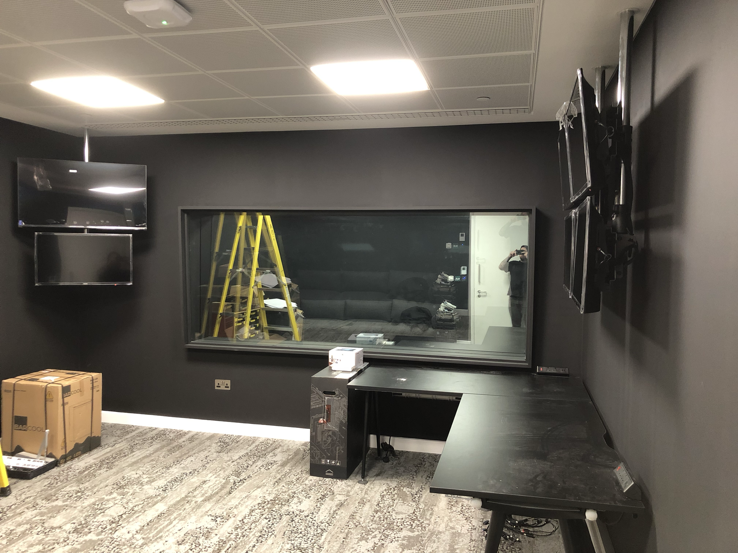 Room with a large window, multiple wall-mounted monitors, a desk, a worker taking a photo, a ladder, boxes, and various electronic equipment.