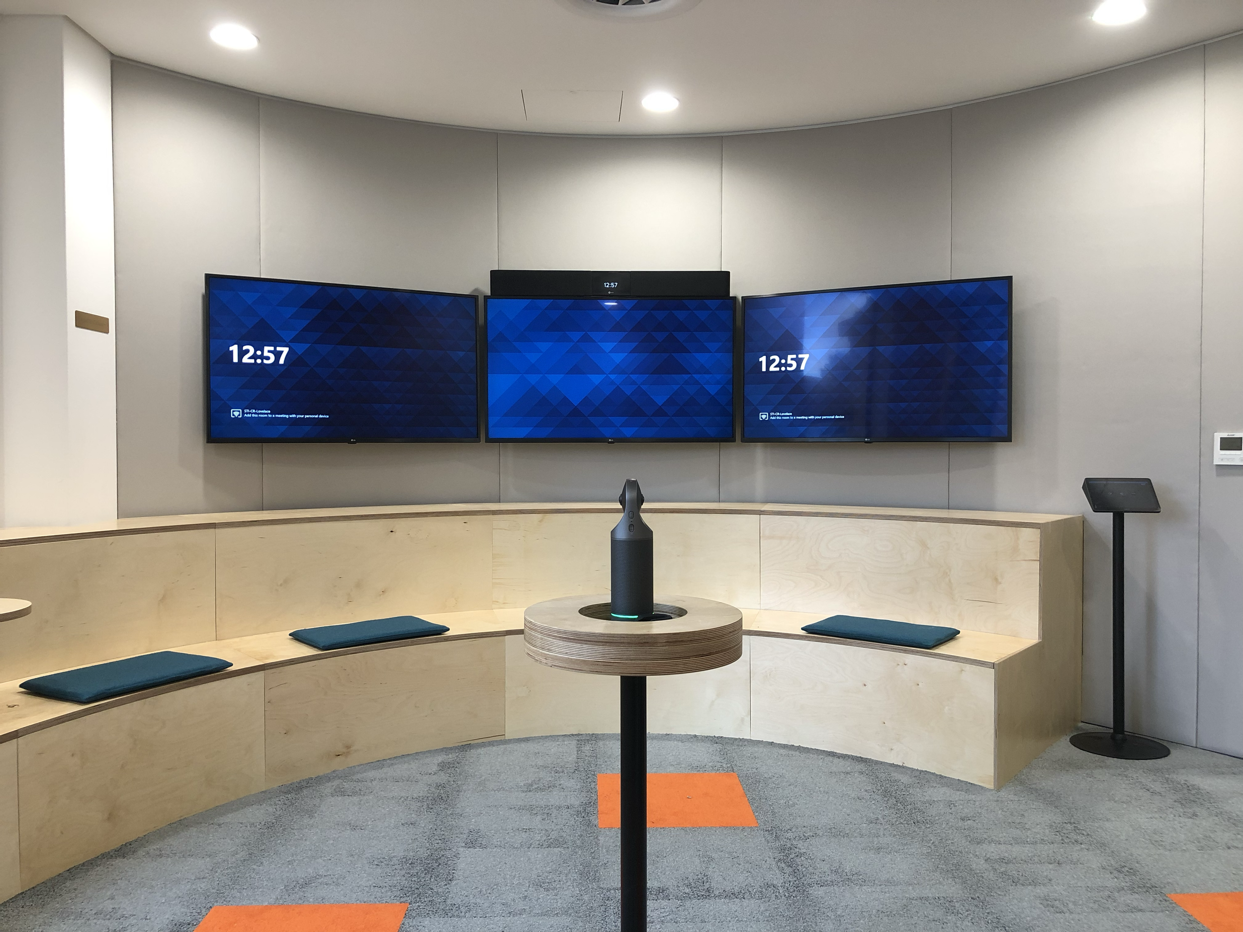 A modern conference room featuring three wall-mounted digital screens displaying the time 12:57. A wooden curved bench with blue cushions faces the screens, with a small table in front holding a black speaker or device. There is a stand with a tablet