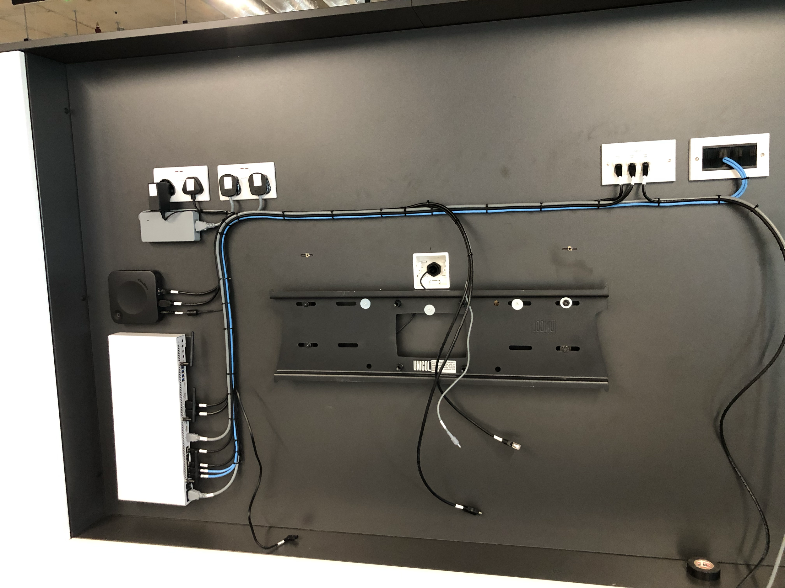 Electronic equipment mounted on a black panel, including power outlets, Ethernet ports, cables, and a network switch, with some cables hanging loosely.