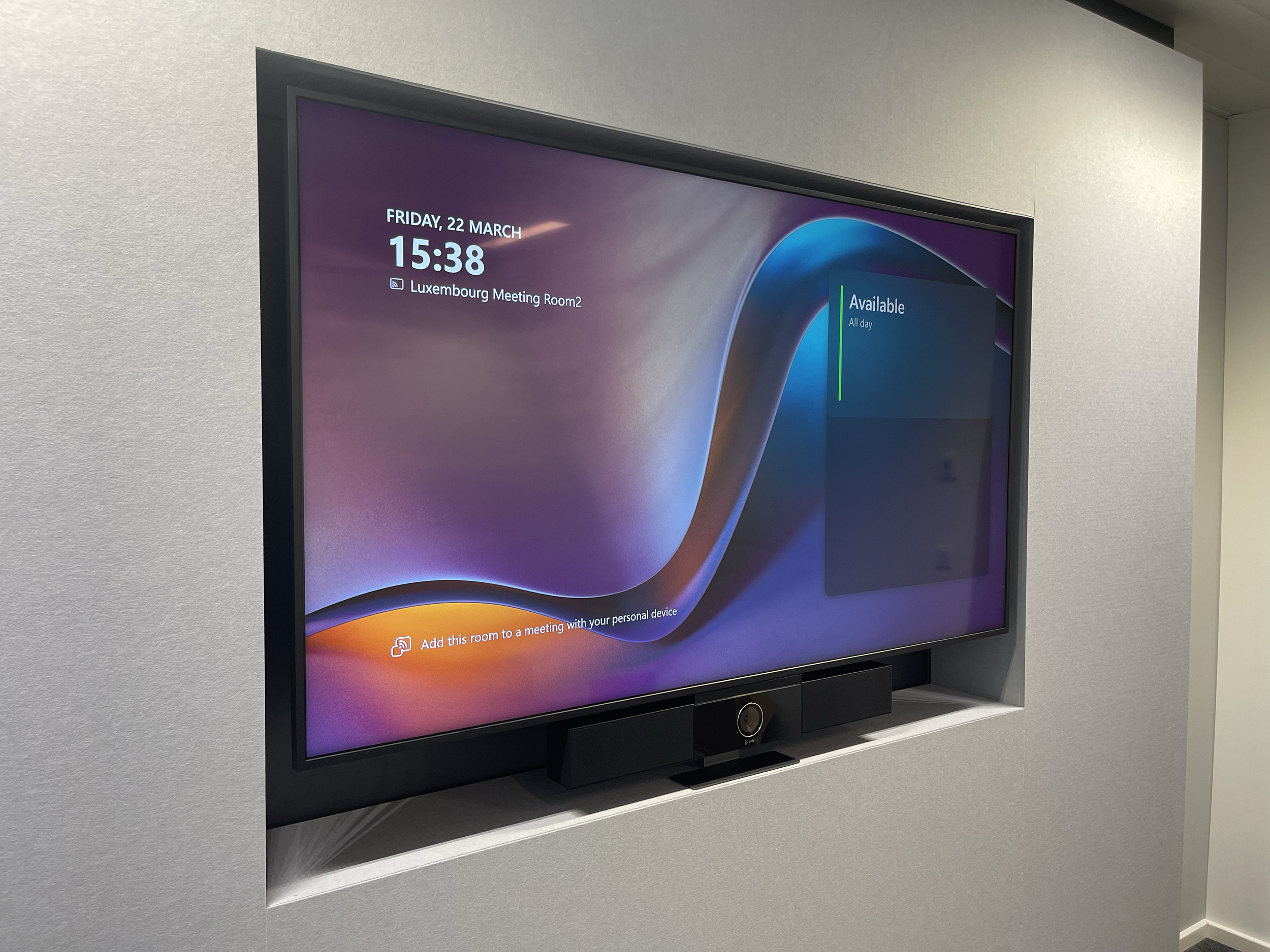 Digital display in a conference room showing the date, time, and room availability, with a speaker and camera below the screen.