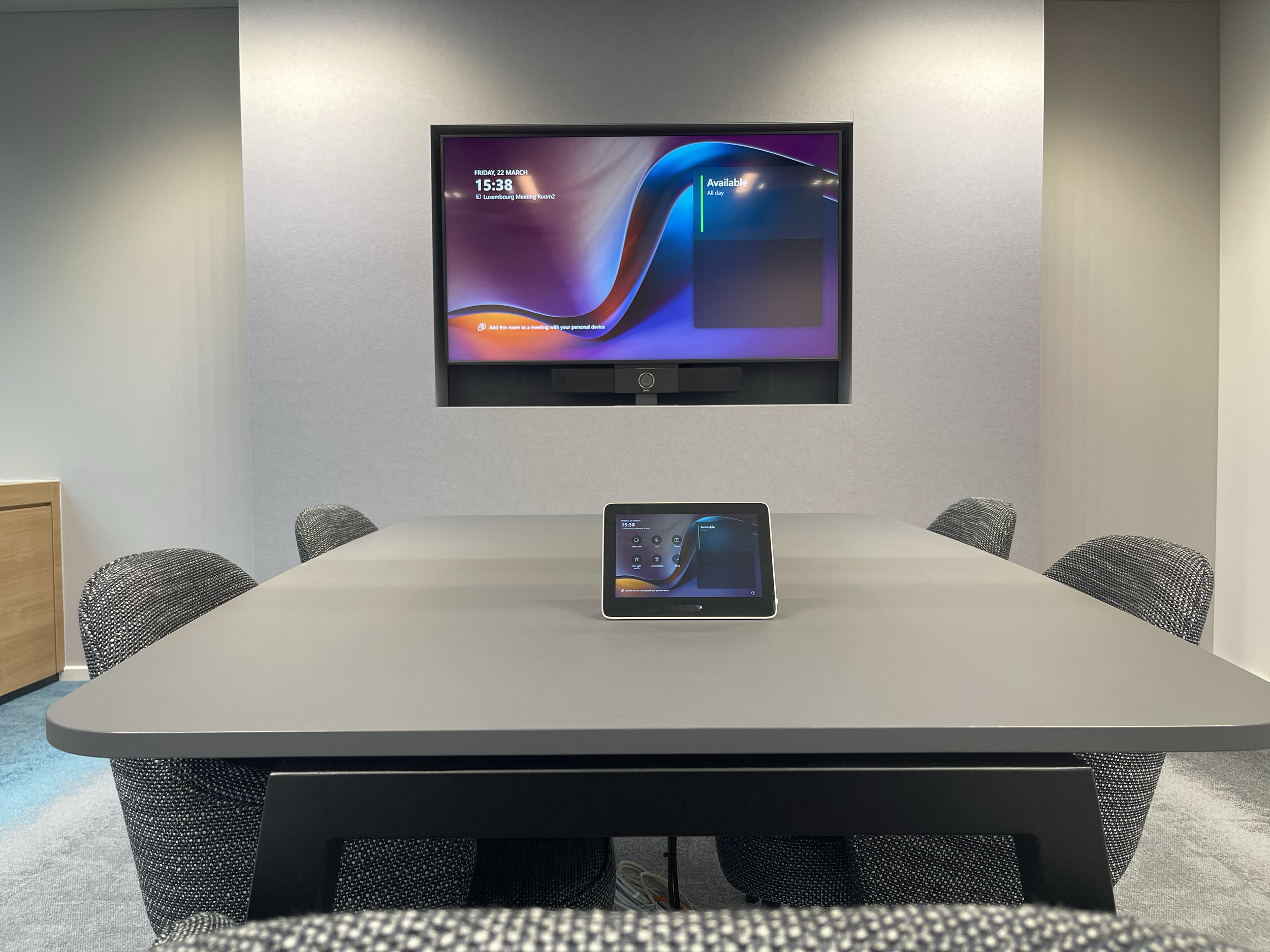 Conference room with a gray table and six black patterned chairs, a wall-mounted screen, and a tablet device on the table.