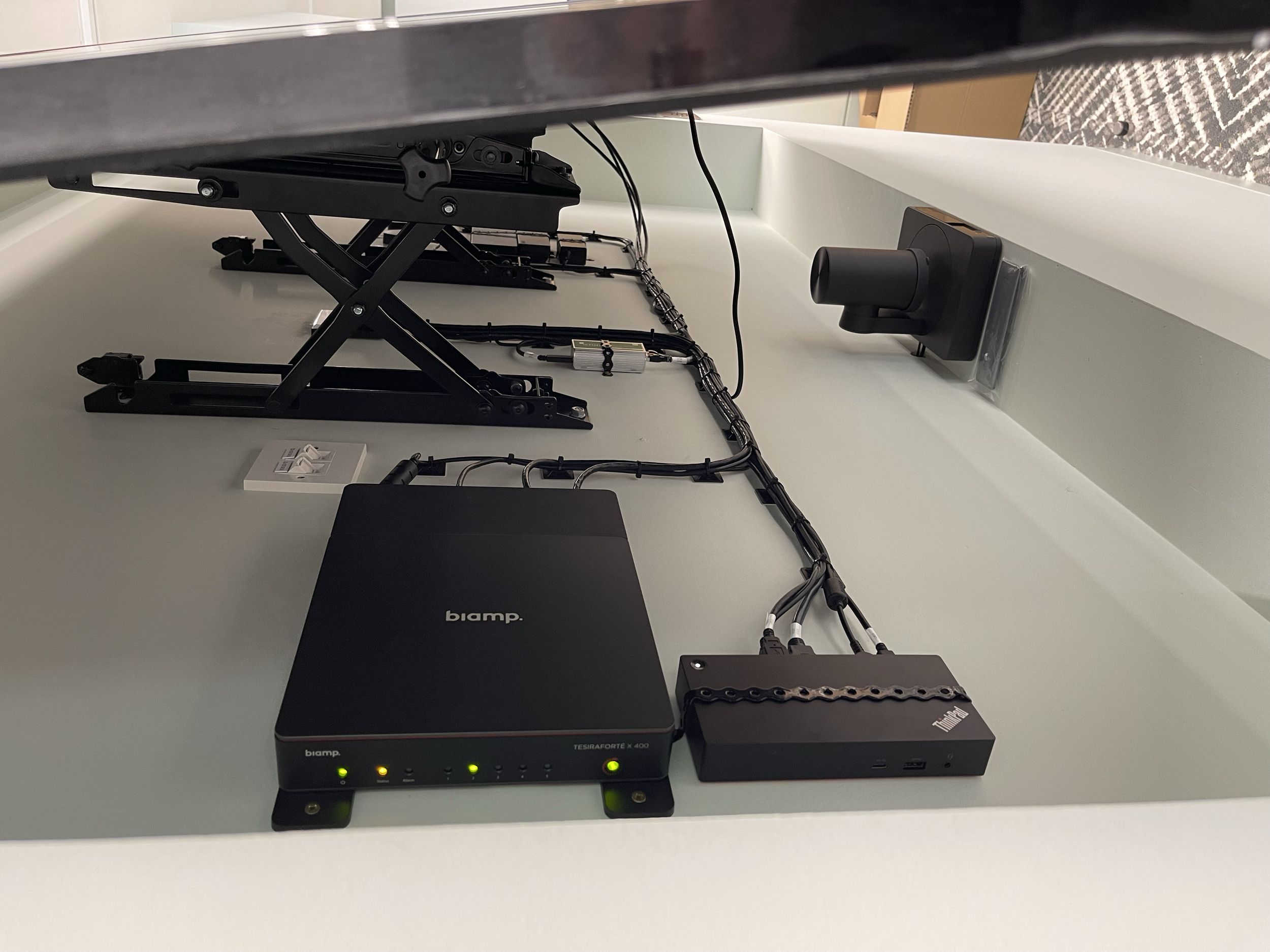 Computer hardware setup including a black biamp device, a black Lenovo ThinkPad docking station, a monitor mount, and various cables on a white desk.