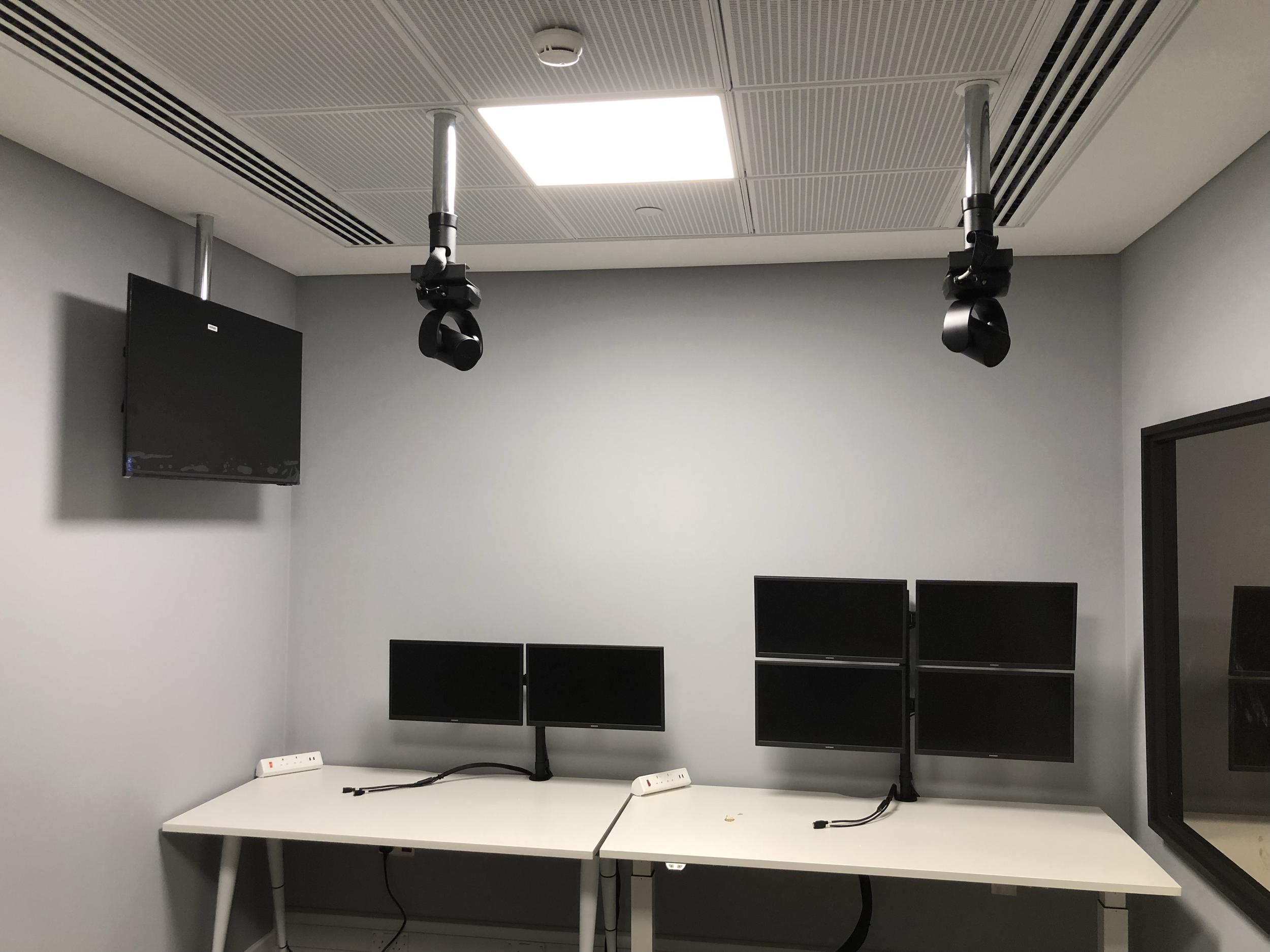 Empty control room with two desks, each equipped with two monitors, and a wall-mounted TV screen. Two ceiling-mounted camera or sensor devices are hanging from the ceiling. The room has a gray color scheme with a modern design.