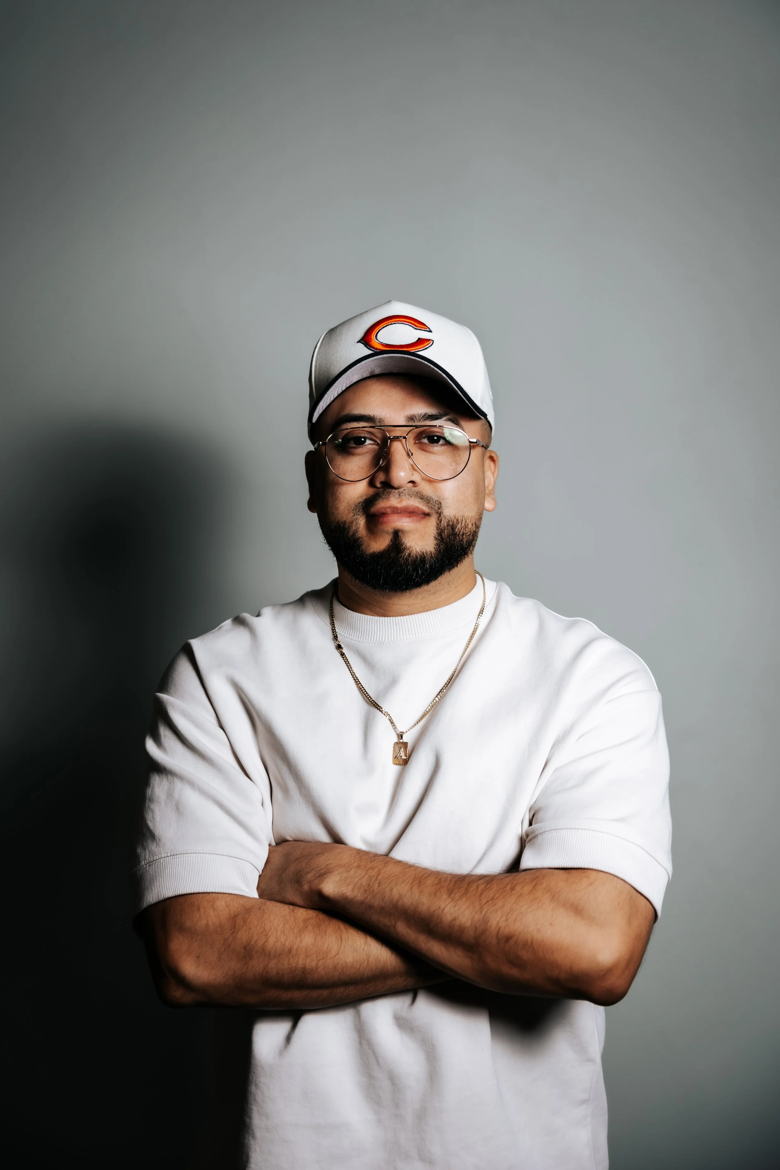 Man with glasses, beard, and mustache wearing a white t-shirt, a gold chain with a pendant, and a white baseball cap with a red and orange sports team logo, standing against a gray background with arms crossed.