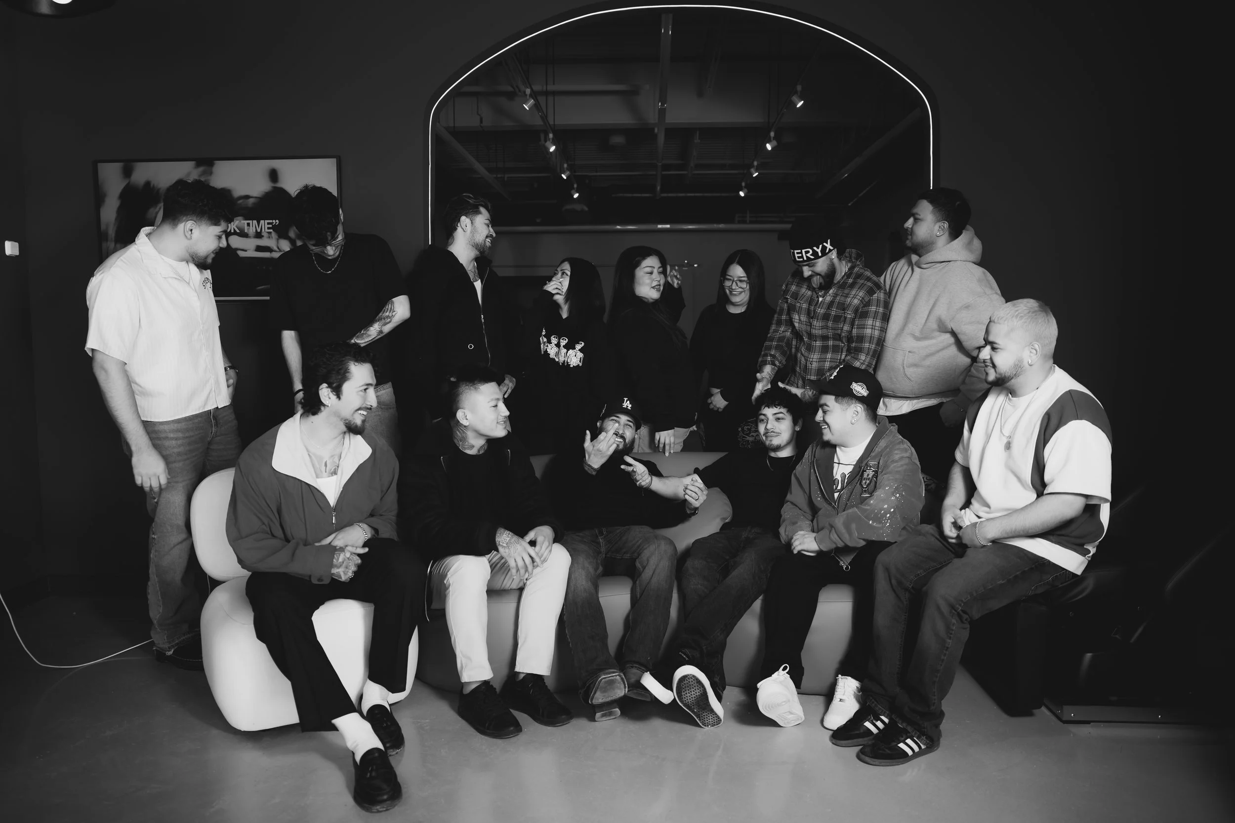 A black-and-white photo of a group of 14 people gathered in a room, some sitting on a sofa and others standing behind. They are smiling and engaging with each other, creating a lively social atmosphere.