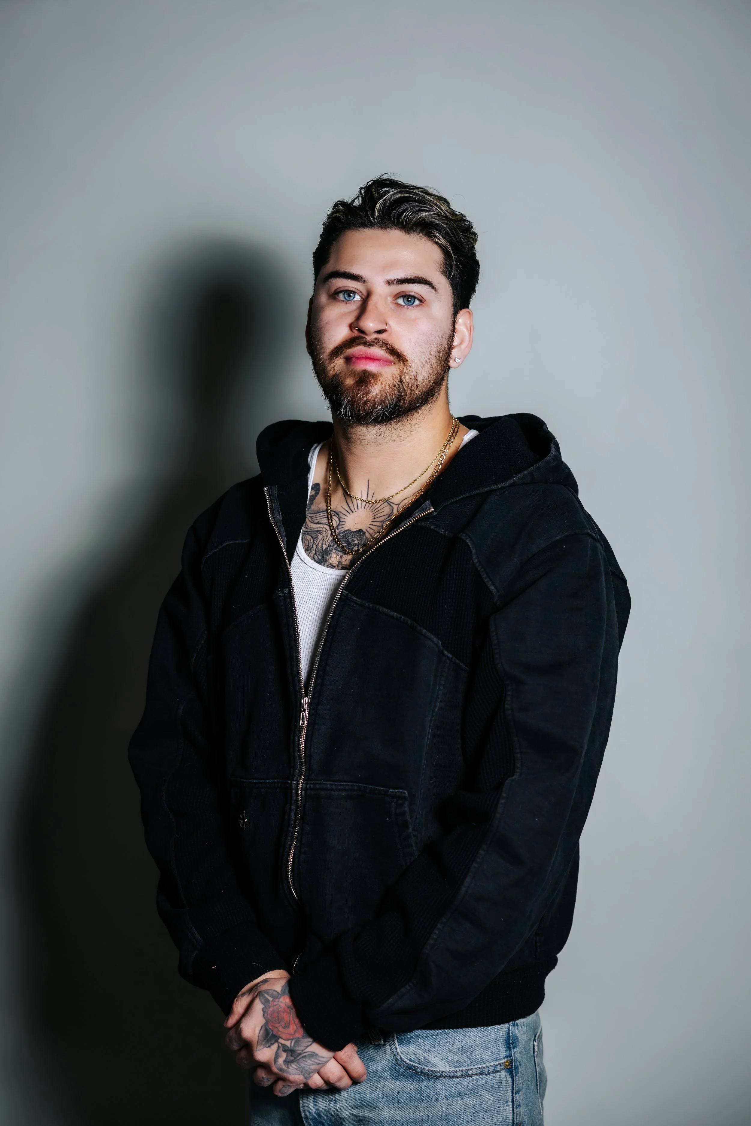 Portrait of a young man with tattoos, beard, and blue eyes, wearing a black hoodie and light jeans, standing against a plain wall.