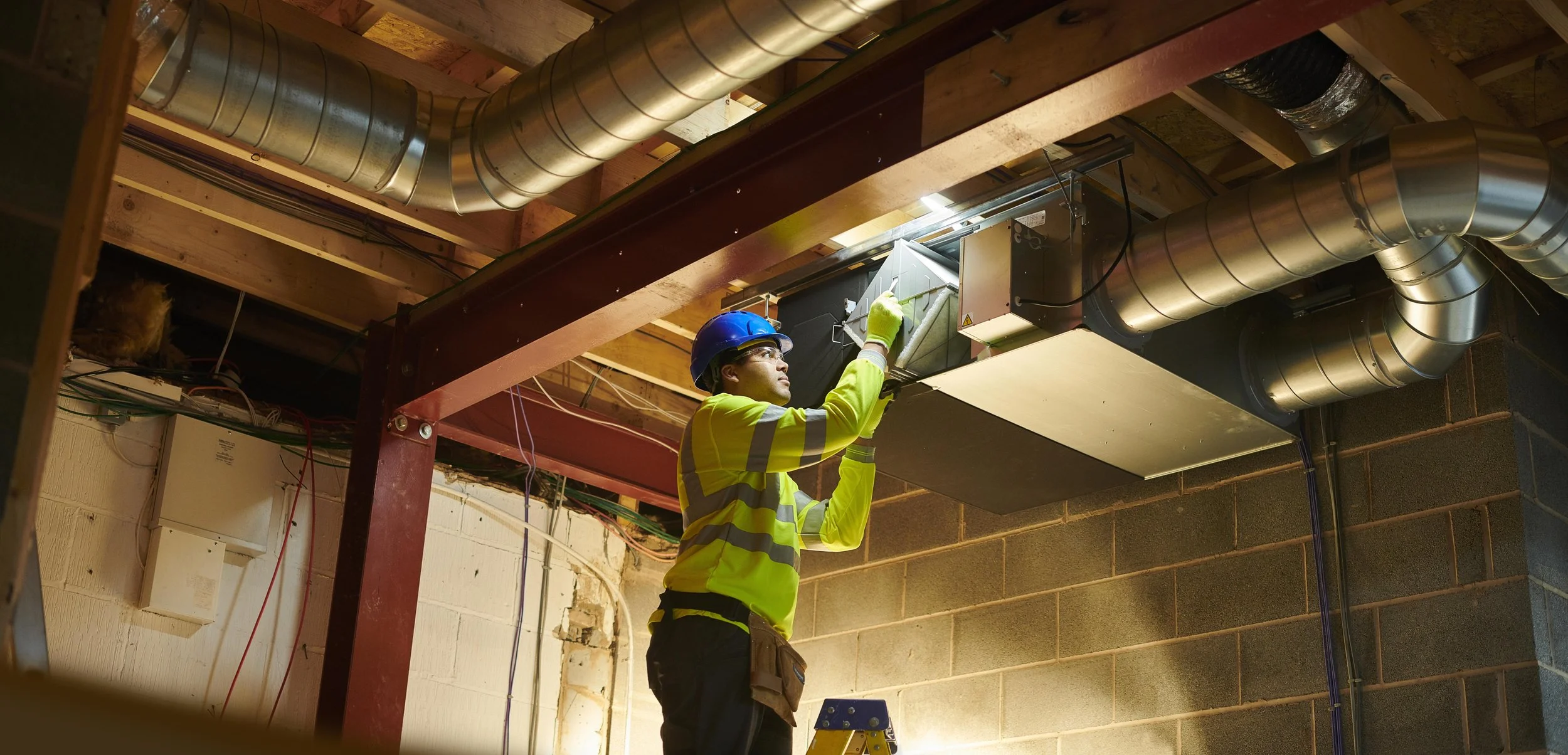 A construction worker wearing a yellow safety vest, blue hard hat, glasses, and gloves inspects and works on a large HVAC duct system installed in an unfinished building with exposed wooden beams and brick walls.