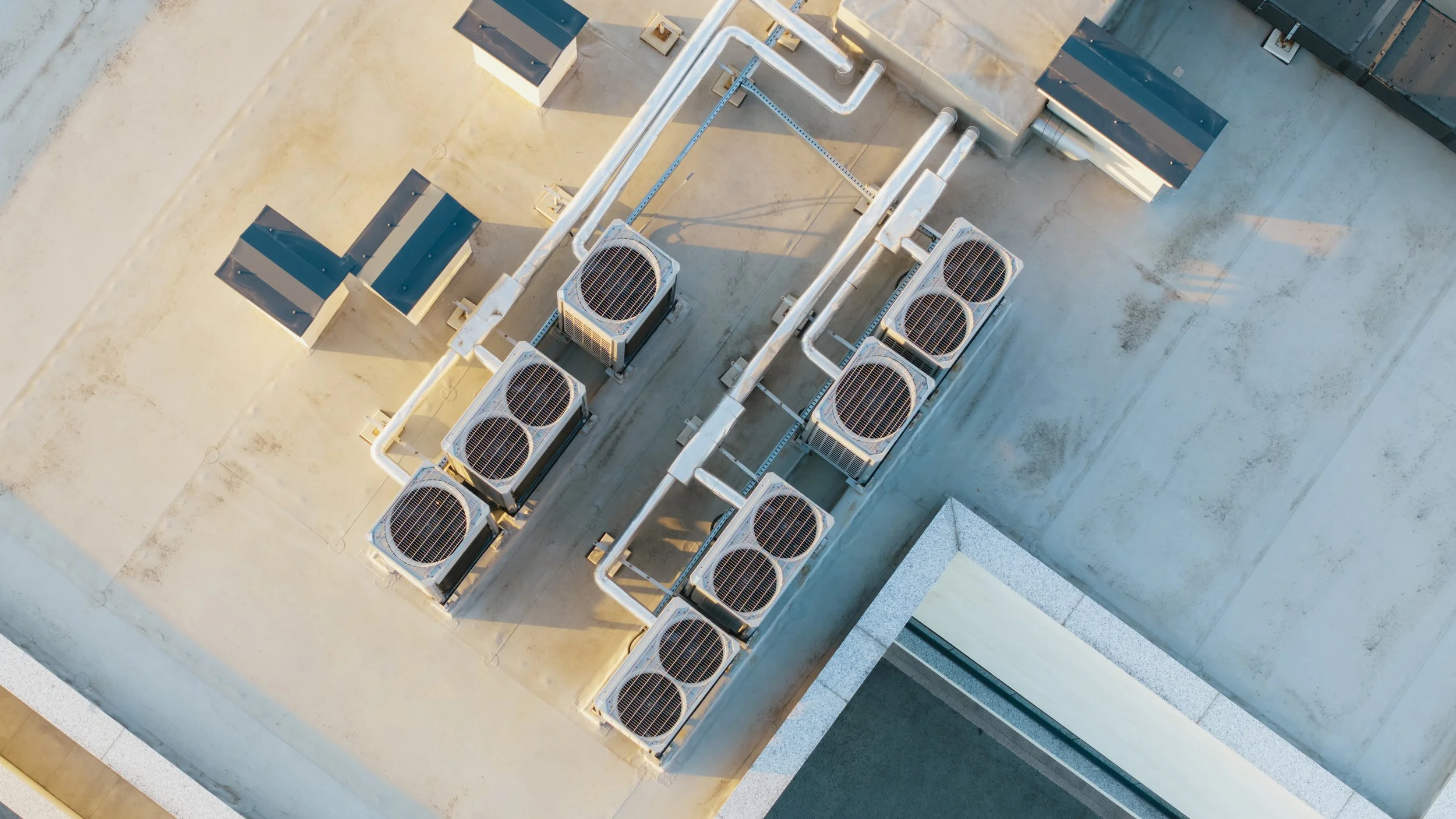 Top-down view of an array of air conditioning units on a building rooftop with visible pipes.