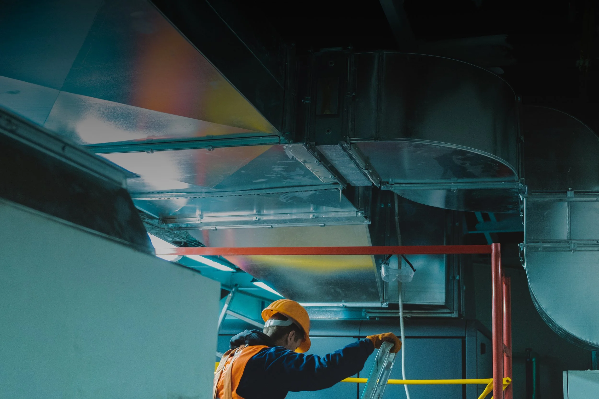 Worker in orange safety gear inspecting ventilation ducts in an industrial setting.
