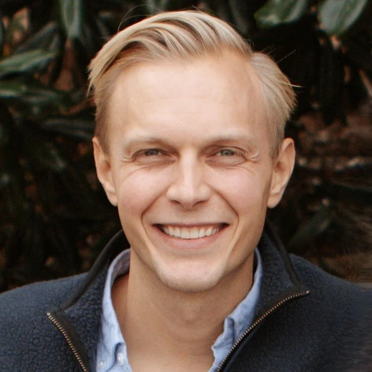 A young man with blonde hair and blue eyes smiling outdoors, wearing a dark jacket and a light blue collared shirt.