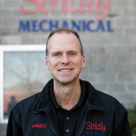A man with short hair and a friendly smile standing outdoors in front of a sign that reads 'Strictly Mechanical' in red and blue letters.