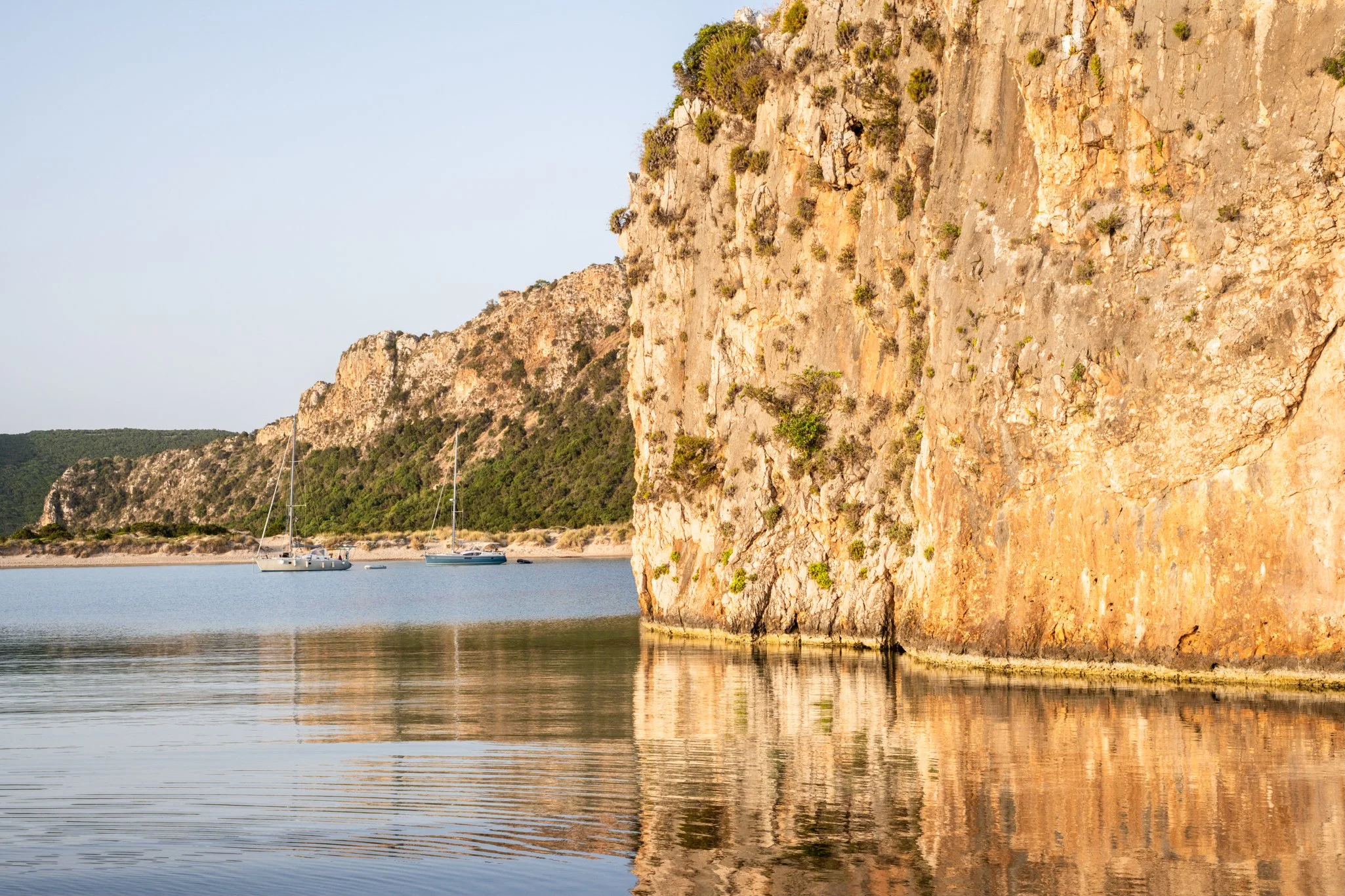 Wall art photography of calm water reflecting sunlit cliffs at Voidokilia Beach in Messinia, Greece with anchored boats in the distance.