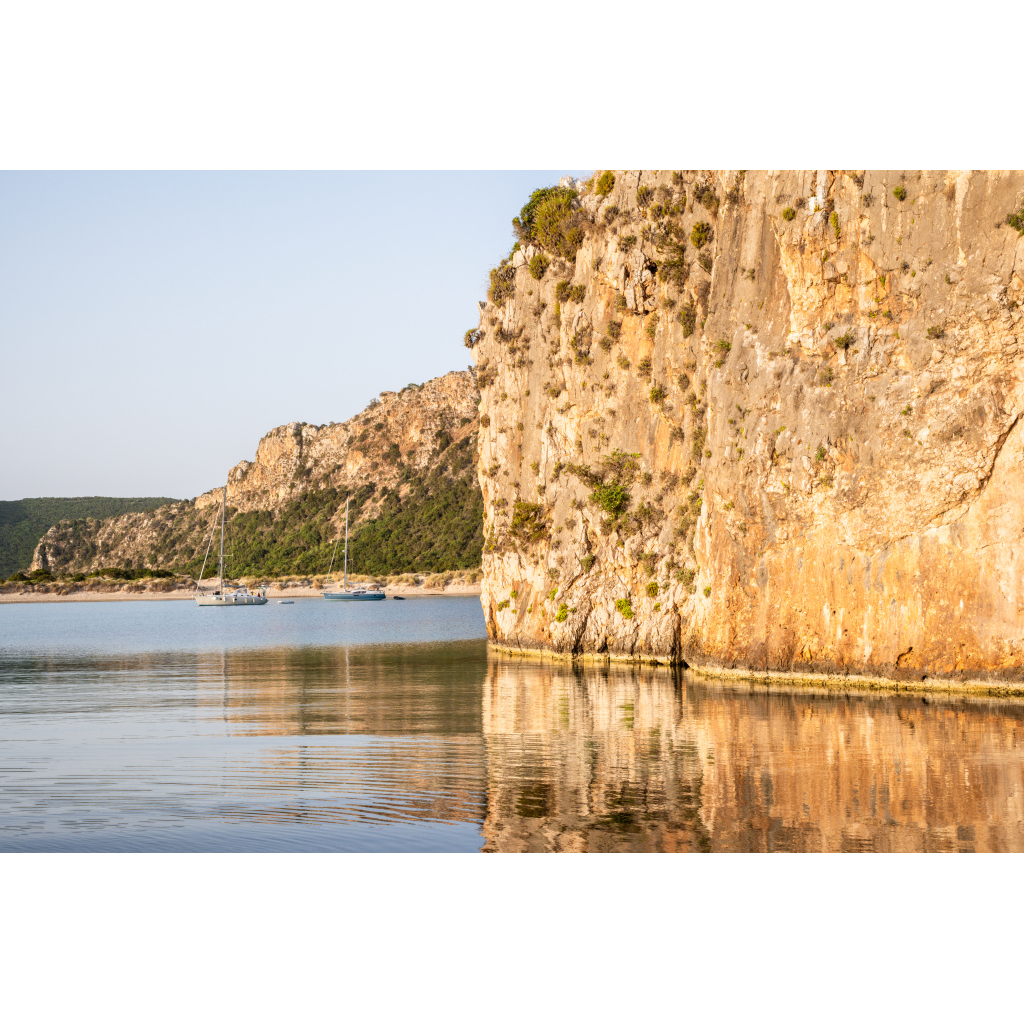 Panoramic view of calm water reflecting sunlit cliffs at Voidokilia Beach in Messinia, Greece with anchored boats in the distance.