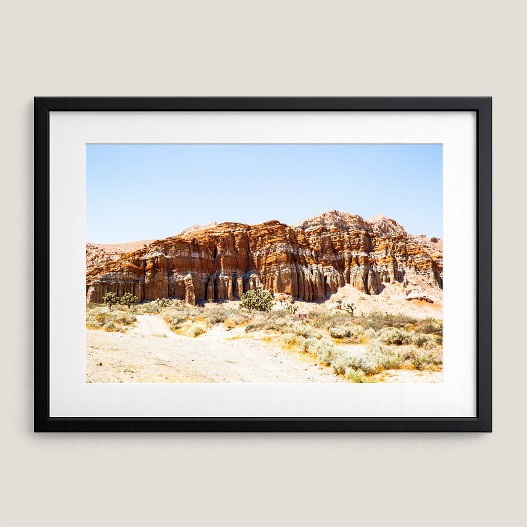 Framed Southwest desert landscape print with black frame and white mat, featuring towering red rock cliff formations.