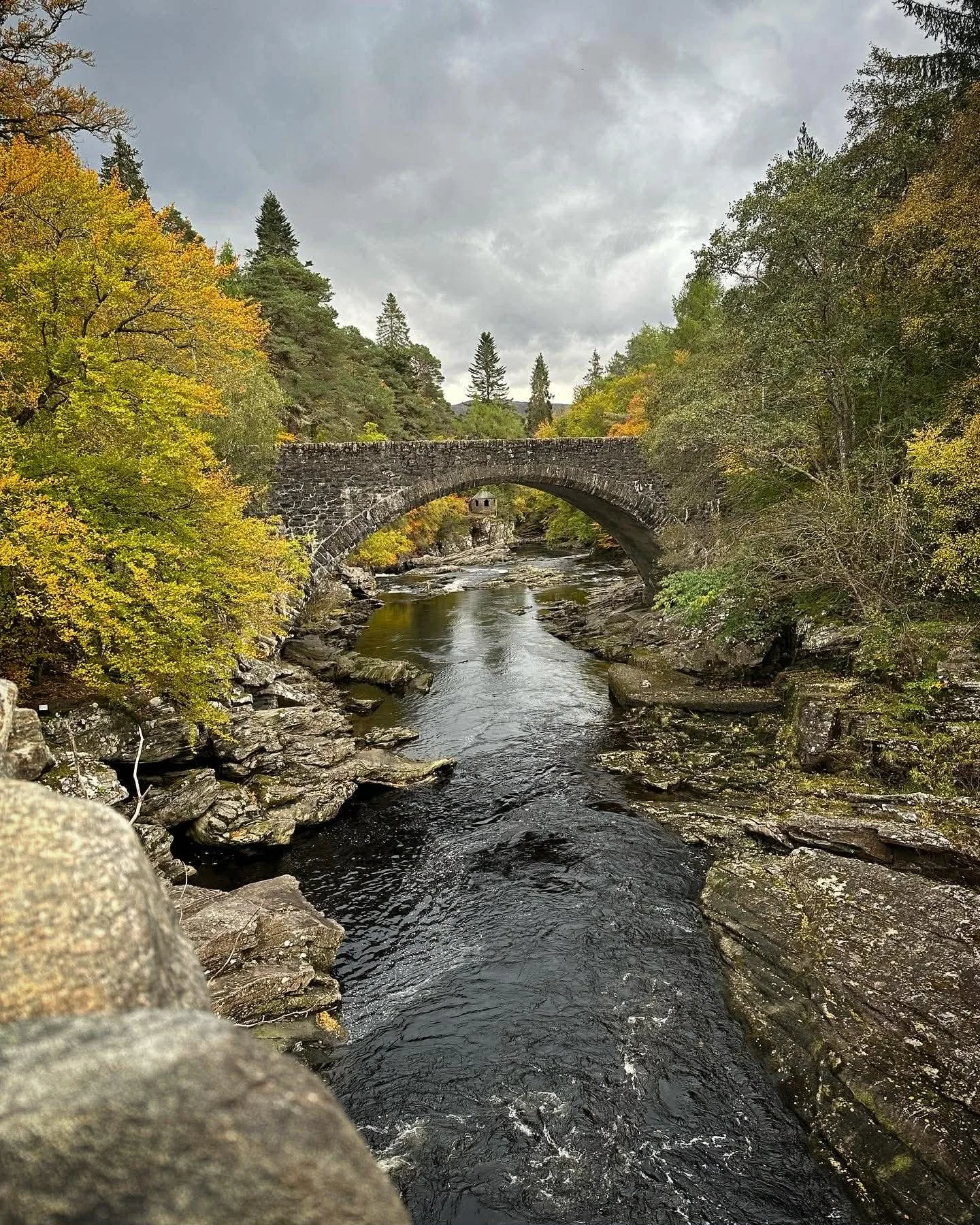 Scenic view of a stone arch bridge over a rocky river with autumn-colored trees on both sides and a cloudy sky above.