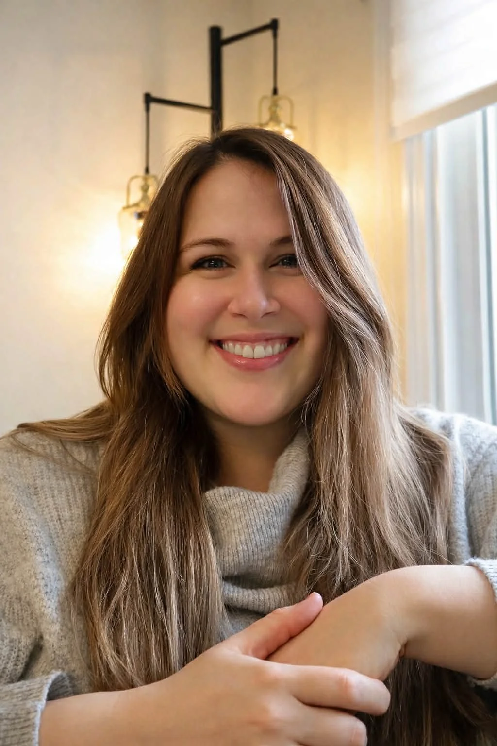 A young woman with long brown hair smiling at the camera, sitting indoors near a window and decorative wall lights.