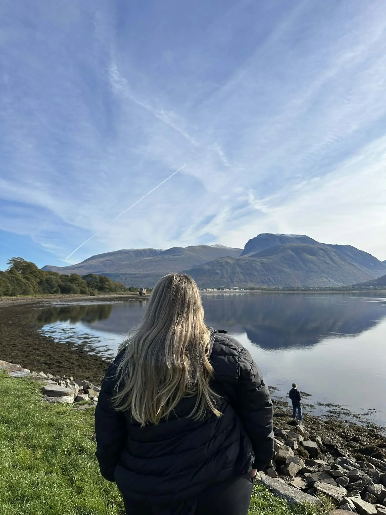 A woman with long blonde hair wearing a black jacket gazes out at a calm lake with green shoreline and mountain range in the background, with a person standing near the water's edge.