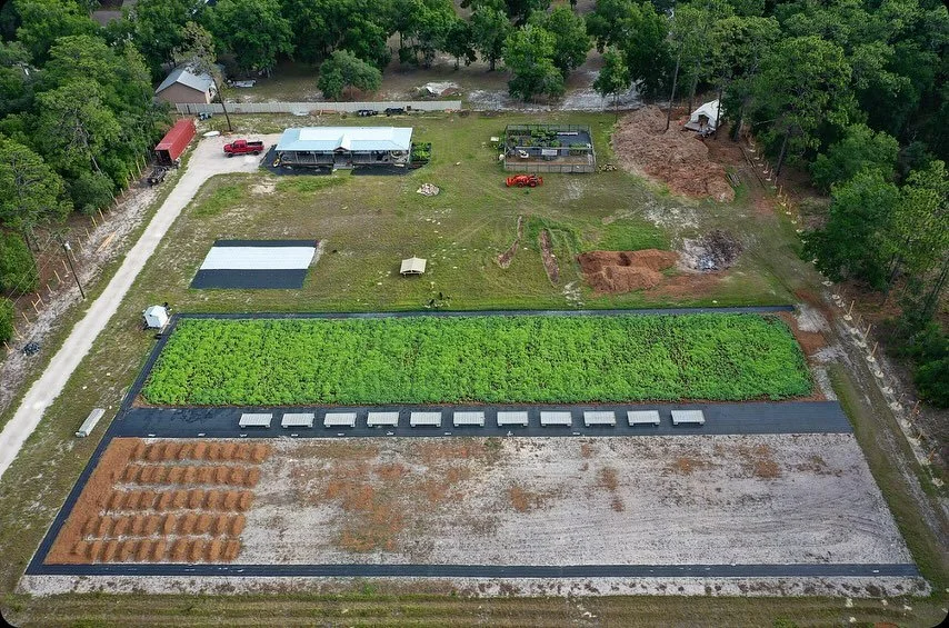 Aerial view of a farm with a large green vegetable garden, smaller plots of soil, and various structures surrounded by trees.