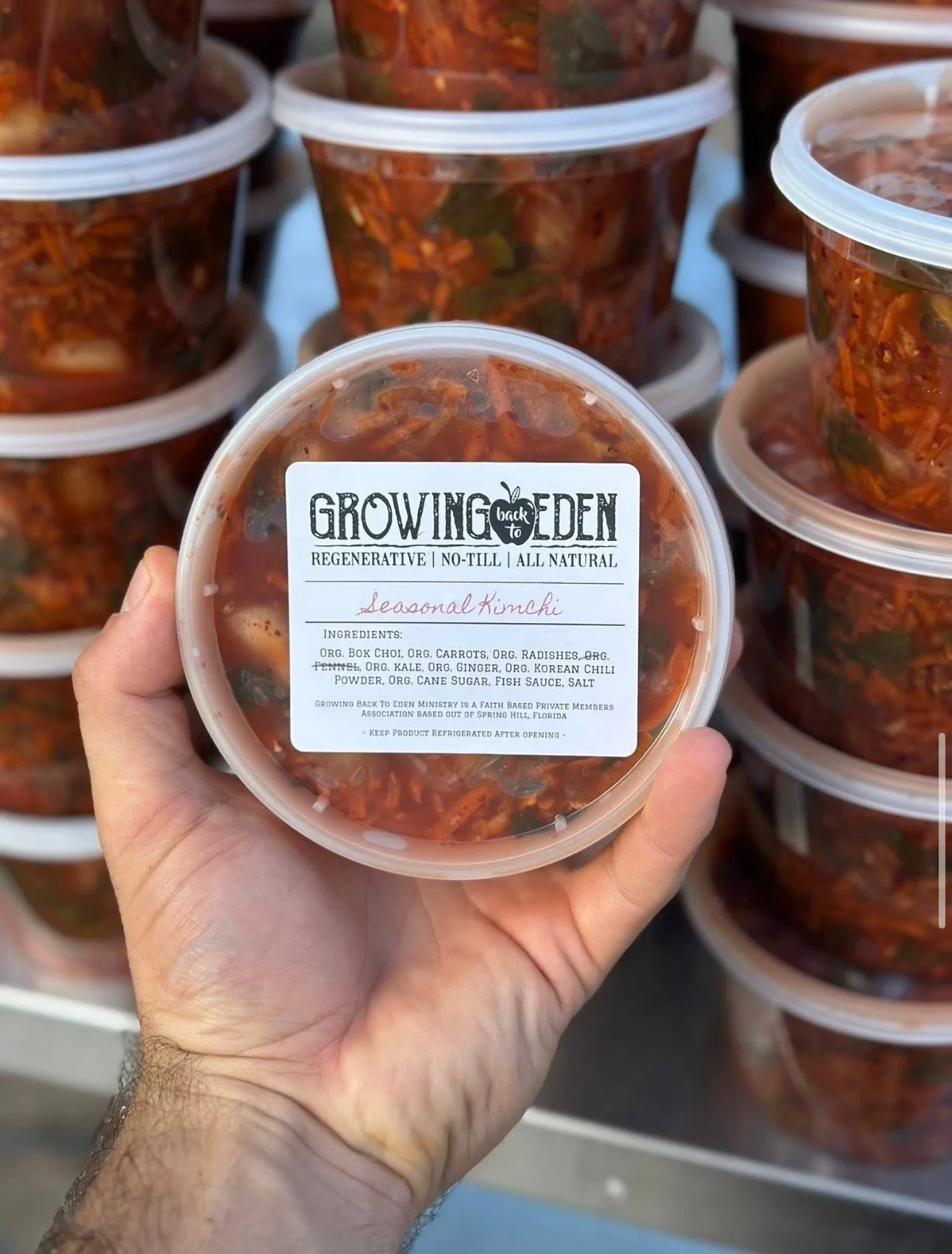 A hand holding a container of seasonal kimchi from Growing Back to Eden, with several more containers of kimchi stacked in the background.