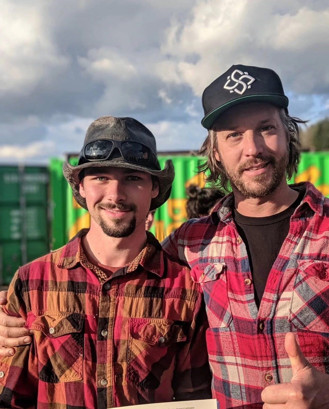 Two men standing outdoors, smiling, with one giving a thumbs up, against a background of green trucks and cloudy sky.