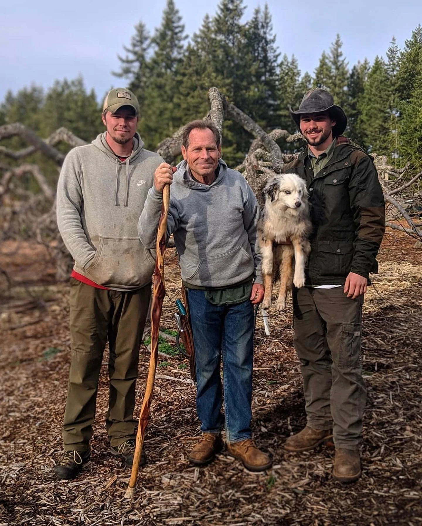 Three men and a dog in a forest, surrounded by fallen trees and green trees in the background. The man in the middle holds a walking stick, and the man on the right is holding a dog.