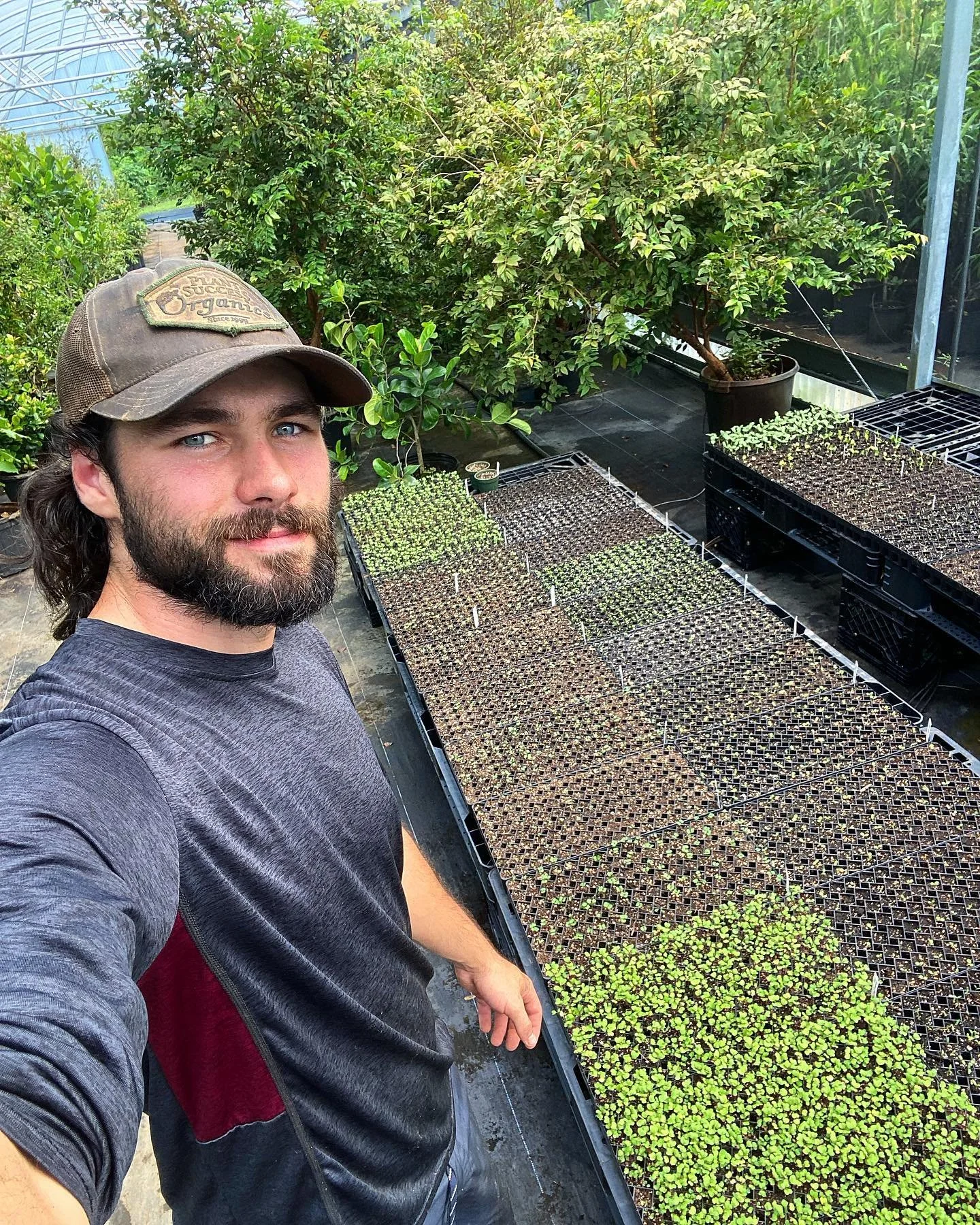 A man taking a selfie in a greenhouse or nursery with trays of seedlings and small plants on tables, surrounded by lush green plants and trees.