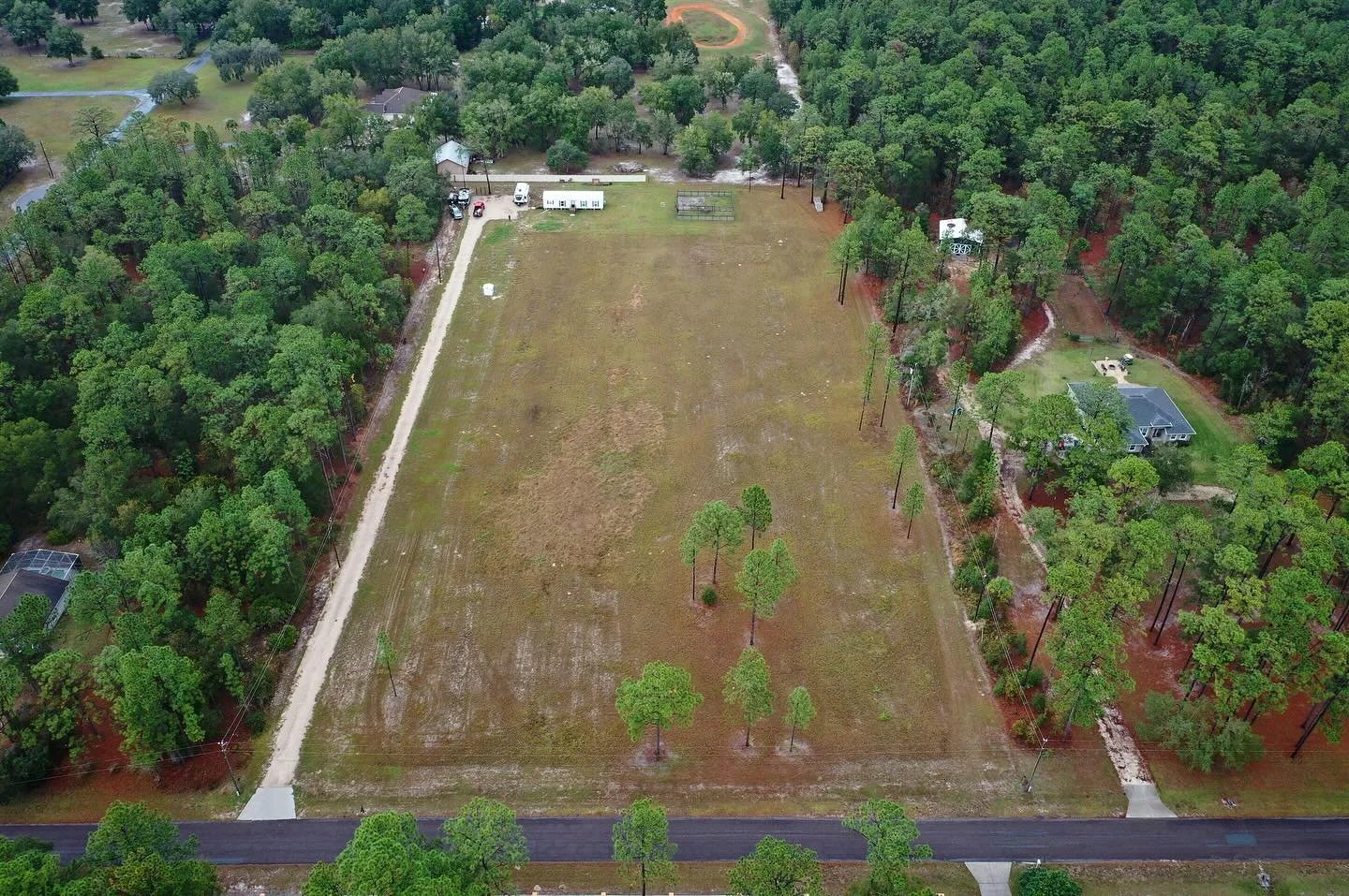 Aerial view of a large open lot surrounded by trees, with a house on the right and a small building at the top, roads, and parked cars.
