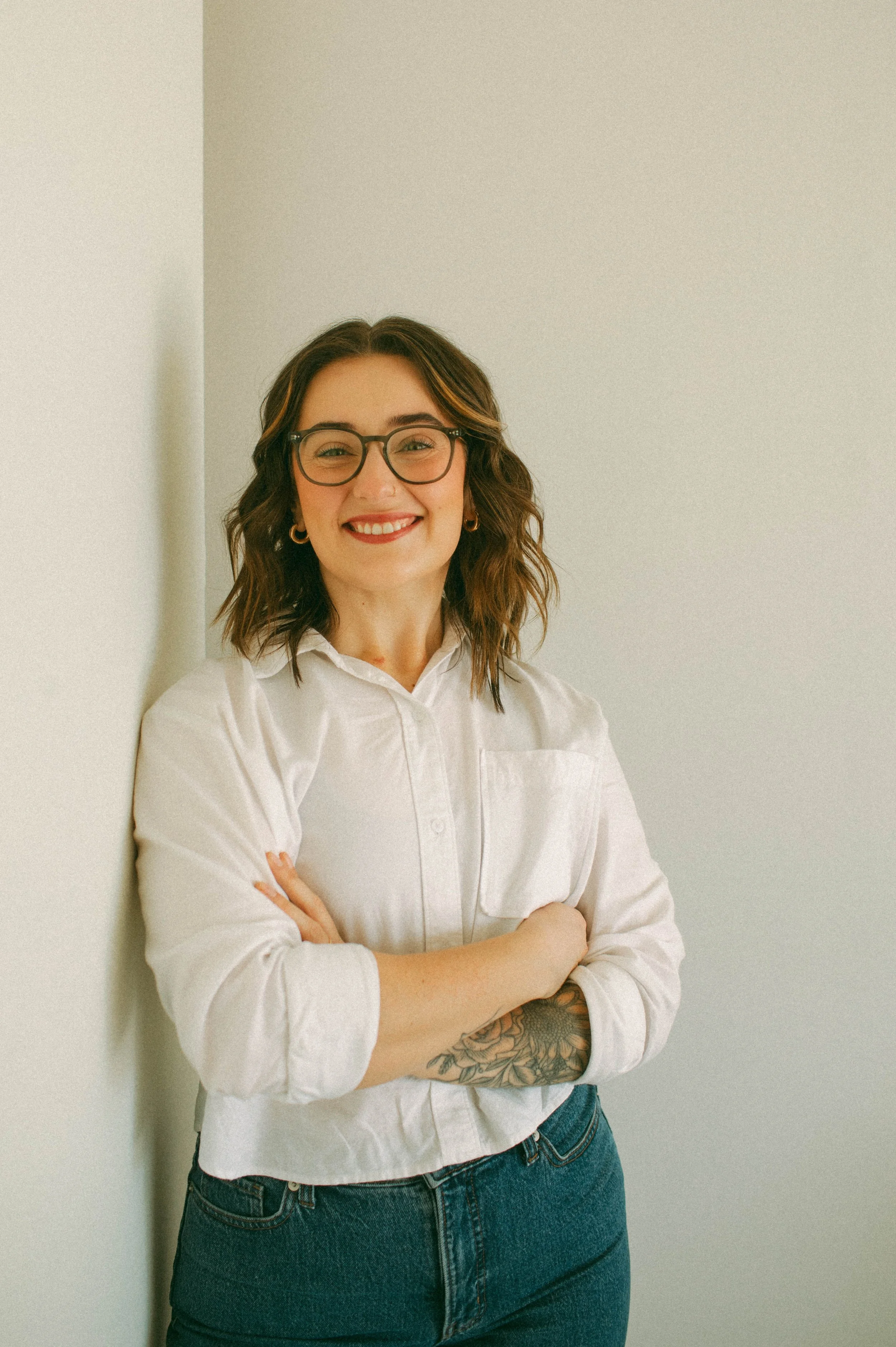 A woman standing with arms crossed, smiling, wearing glasses, a white button-up shirt, and jeans, with a tattoo on her left forearm.
