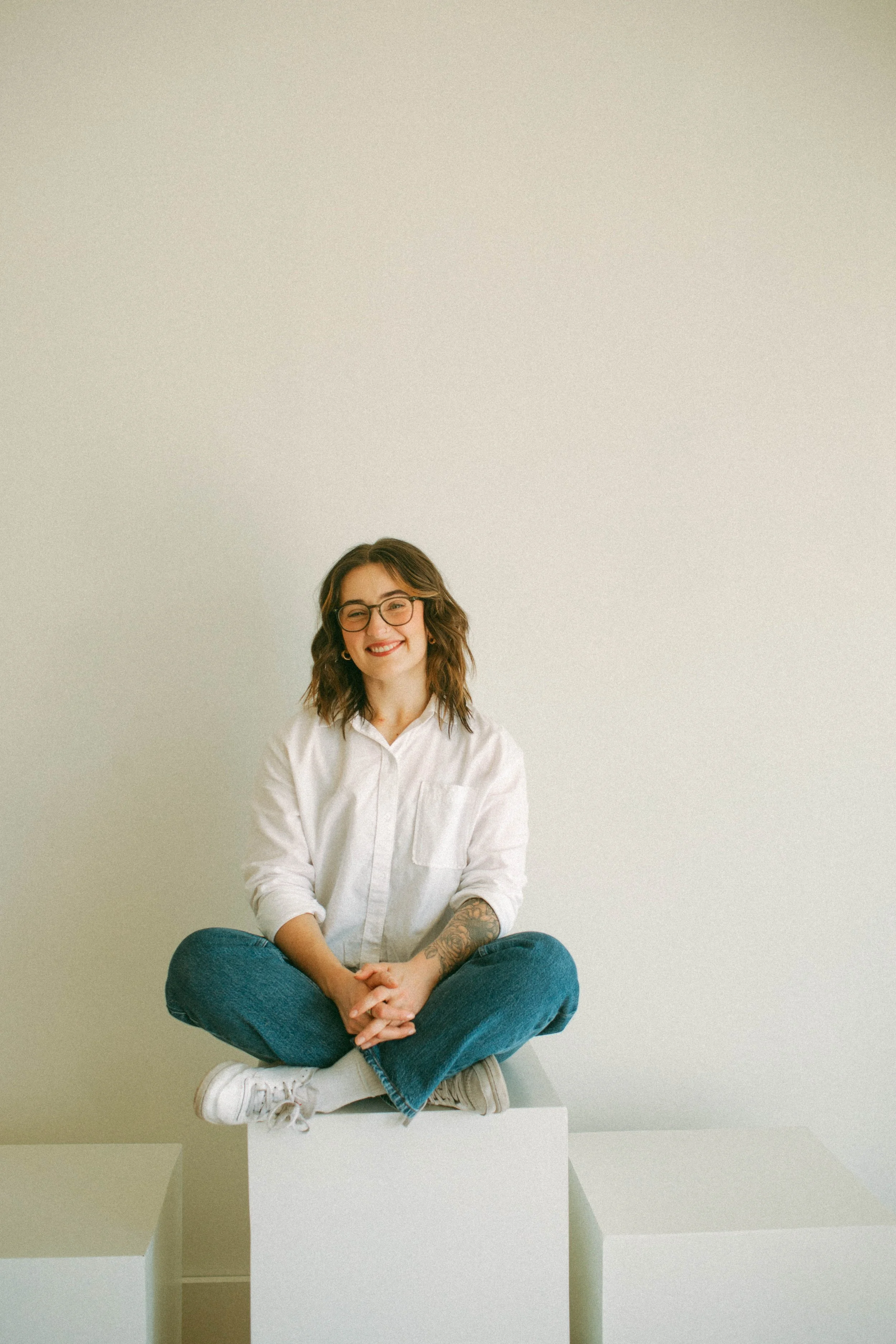 A woman with glasses, smiling, sitting cross-legged on top of white geometric blocks against a plain, light-colored wall.