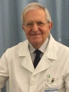 Uomo anziano con capelli grigi, occhiali, vestito da medico con camice bianco e cravatta, sorridente