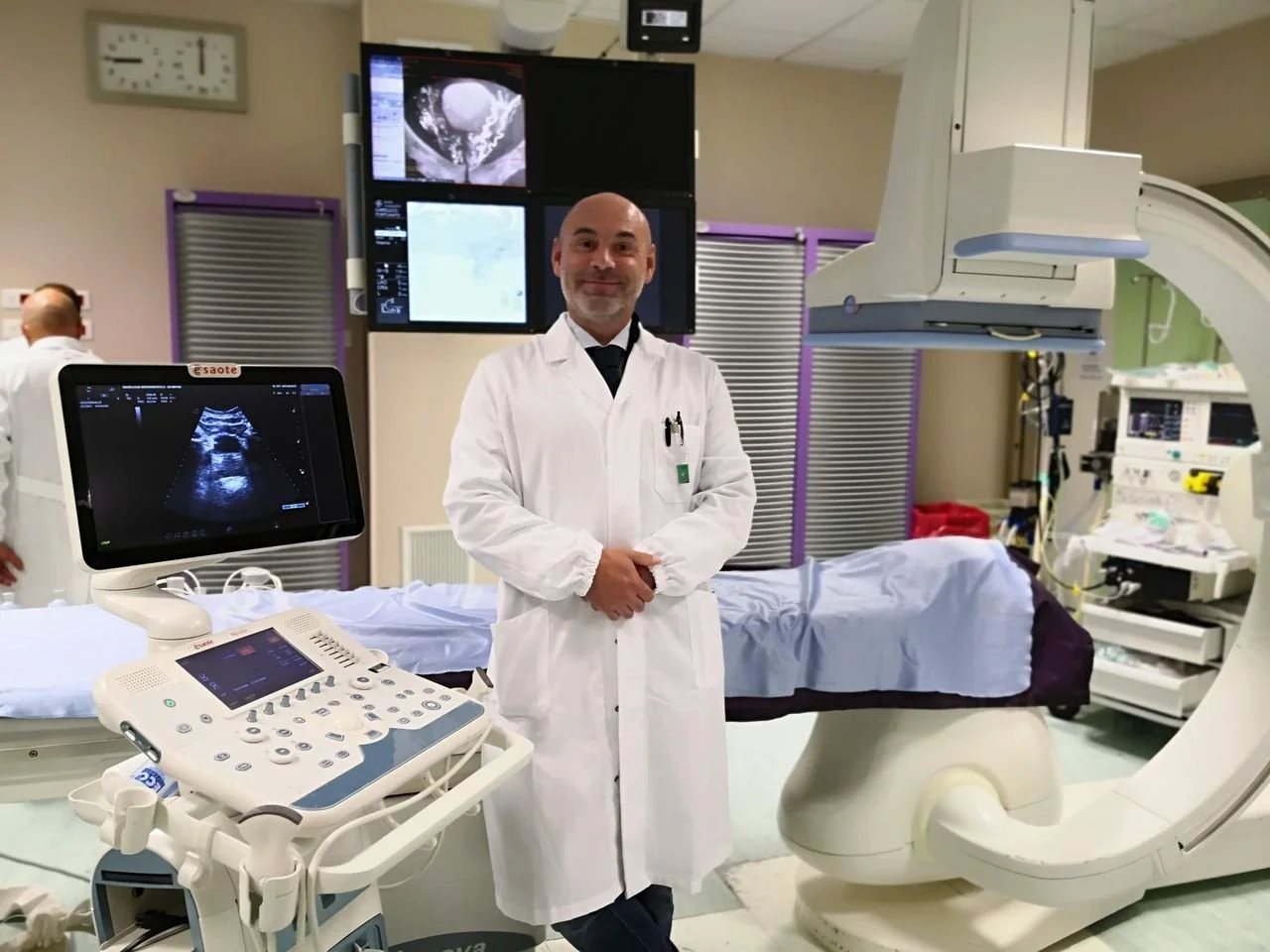 Medico sorridente in camice bianco in stanza con apparecchiature mediche, tra cui macchina per ecografie e monitor, in un ambiente ospedaliero.
