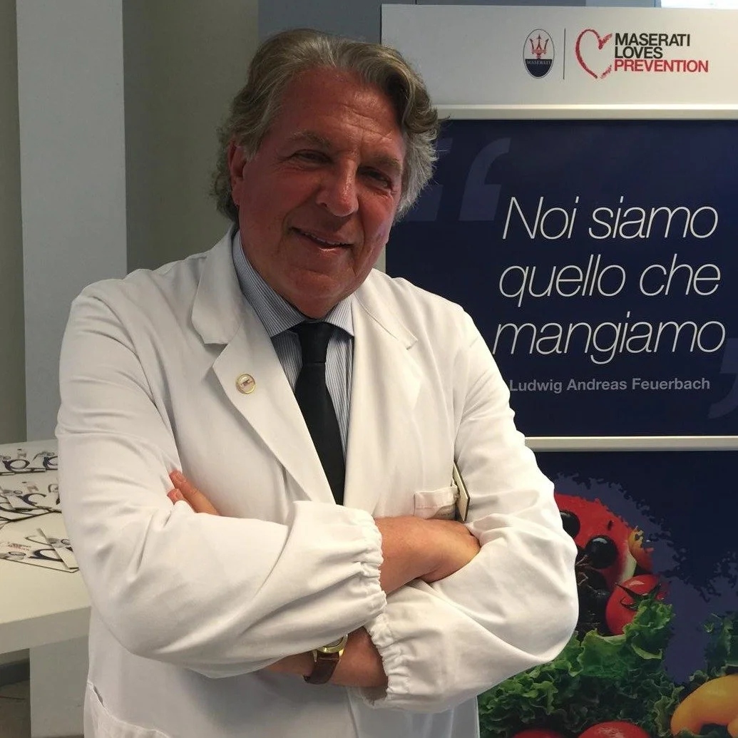Un uomo in camice bianco e cravatta davanti a un poster che promuove la prevenzione contro le malattie, con un messaggio sulla dieta. L'uomo è sorridente con le braccia incrociate.