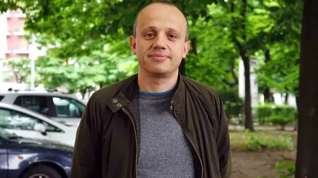Uomo con capelli corti, indossa una giacca nera e un maglione grigio, si trova in un'area verde con alberi e auto in background.