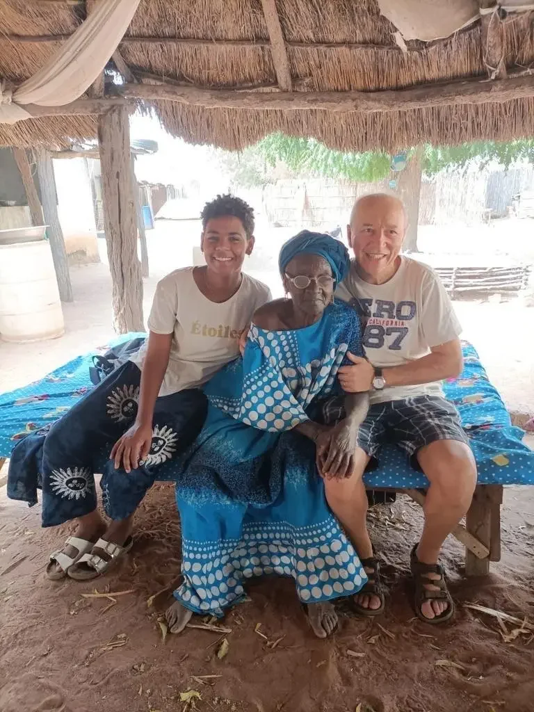 Tre persone sorridenti sedute su una panca sotto una capanna di paglia, con uno sfondo di ambiente rurale. Un'anziana donna indossa abiti tradizionali blu con motivi bianchi, mentre i due uomini e una donna più giovani sono vestiti casual.